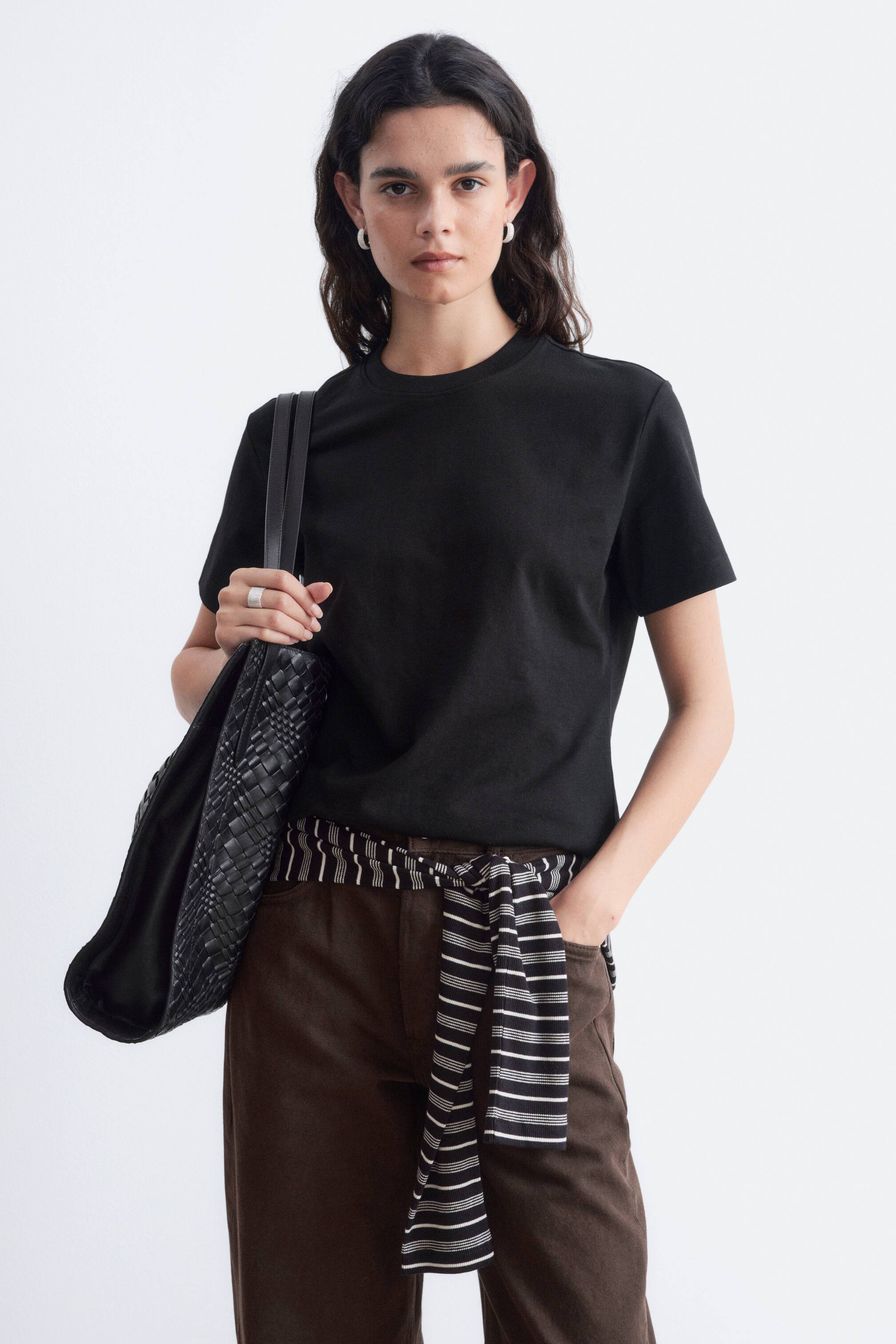 Agrandir l'image: Un T-shirt noir à col rond, à manches courtes et à coupe droite classique est porté par le mannequin. Il est associé à un pantalon marron, un haut rayé noué autour de la taille et un sac tressé noir porté à l’épaule.