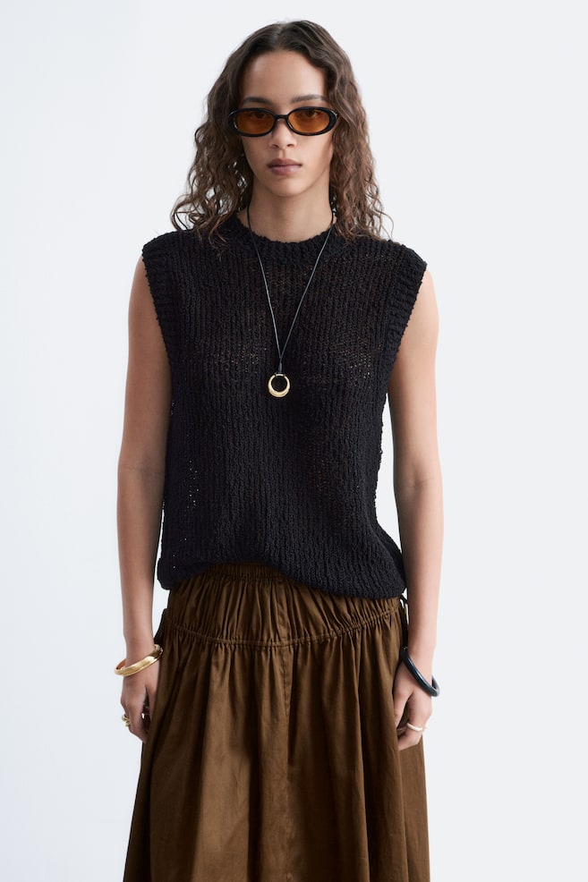 A black open-knit sleeveless top is displayed, featuring a round neckline and a relaxed fit. It is styled over a brown gathered skirt, with a gold ring pendant necklace resting on its front. - 1