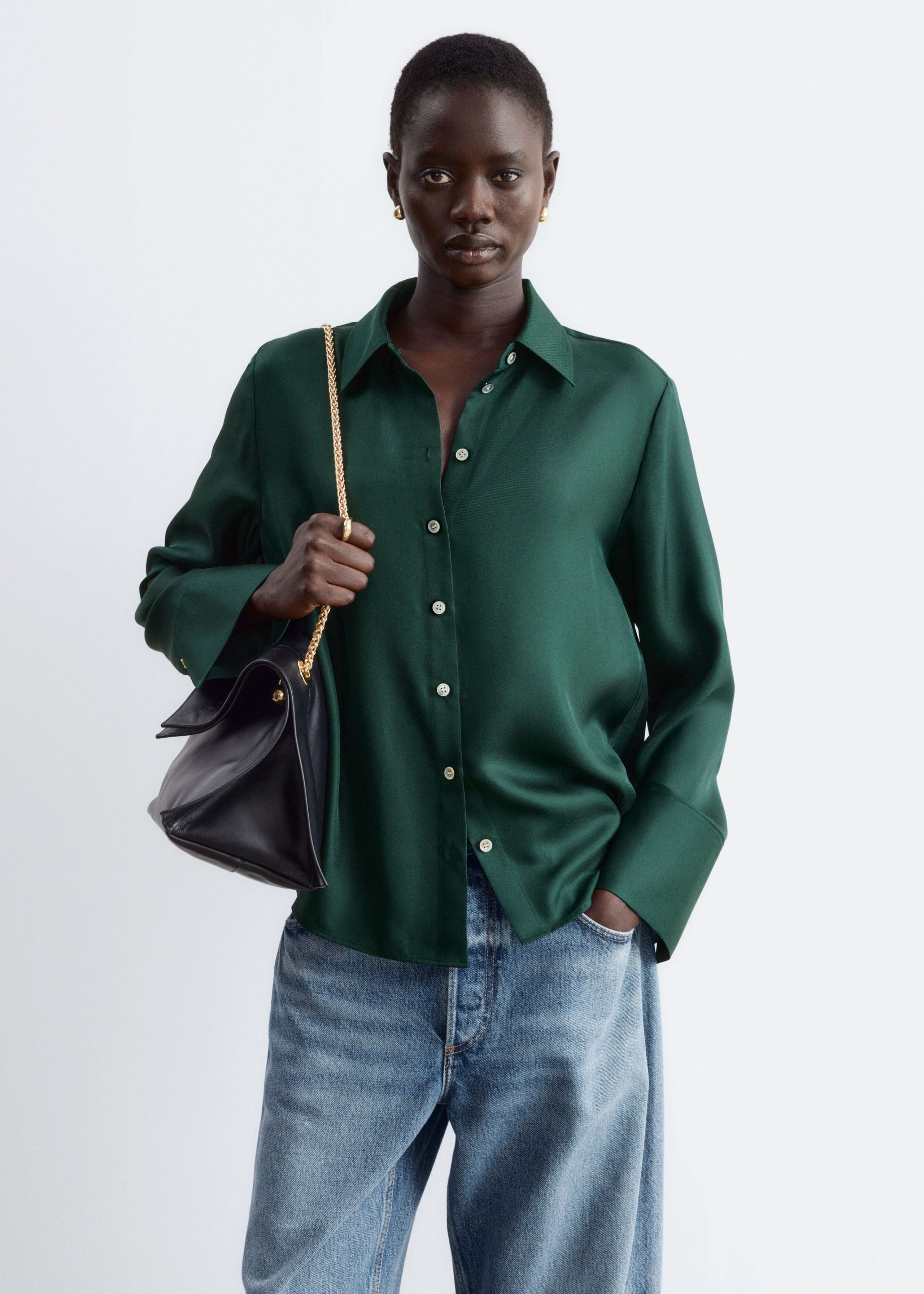 A dark green, long-sleeved silk shirt with a classic collar, button-down front and wide cuffs is being worn by the model. The relaxed-fit shirt is styled with light-wash blue jeans, while the model holds the gold chain strap of a black shoulder bag.
