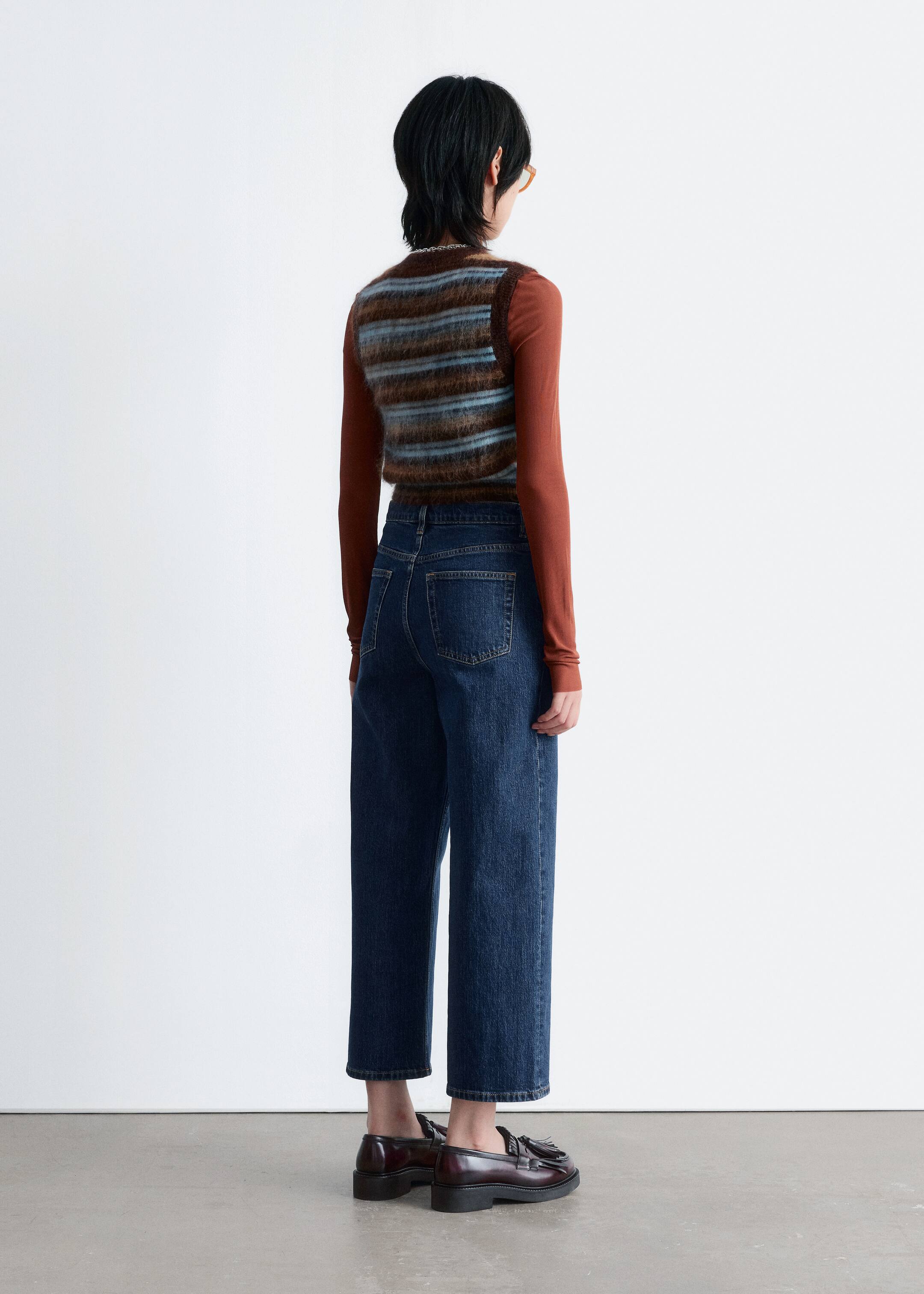 Ver imagen más grande: Desde una vista trasera, se lleva un chaleco de rayas difusas en tonos marrones y azules sobre una camiseta de manga larga color óxido. Se usan jeans de corte recto y lavado oscuro con bolsillos traseros visibles, combinados con mocasines burdeos con borlas.