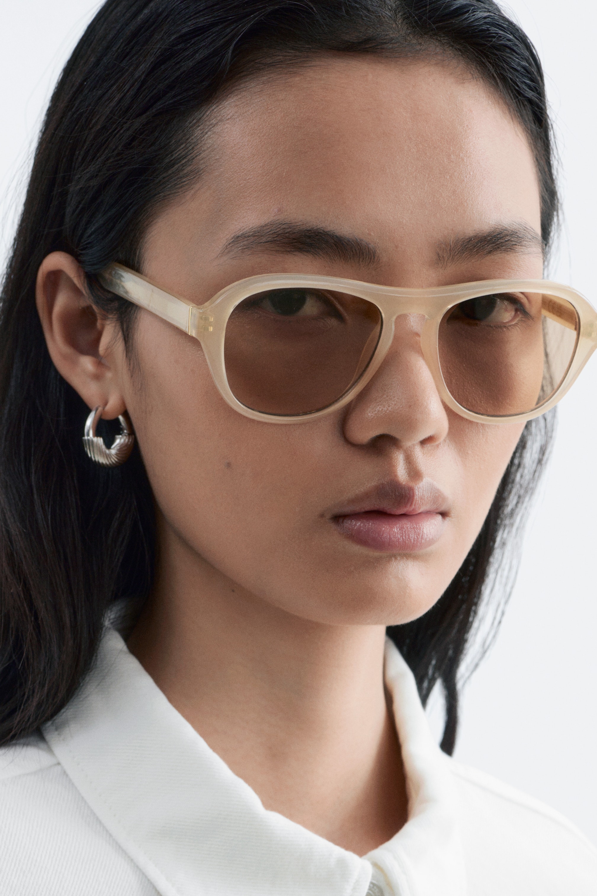 Größeres Bild ansehen: Sonnenbrille mit transparenten, hellbeigen, sanft abgerundeten quadratischen Rahmen und braunen Tönungslinsen wird vom Model getragen.