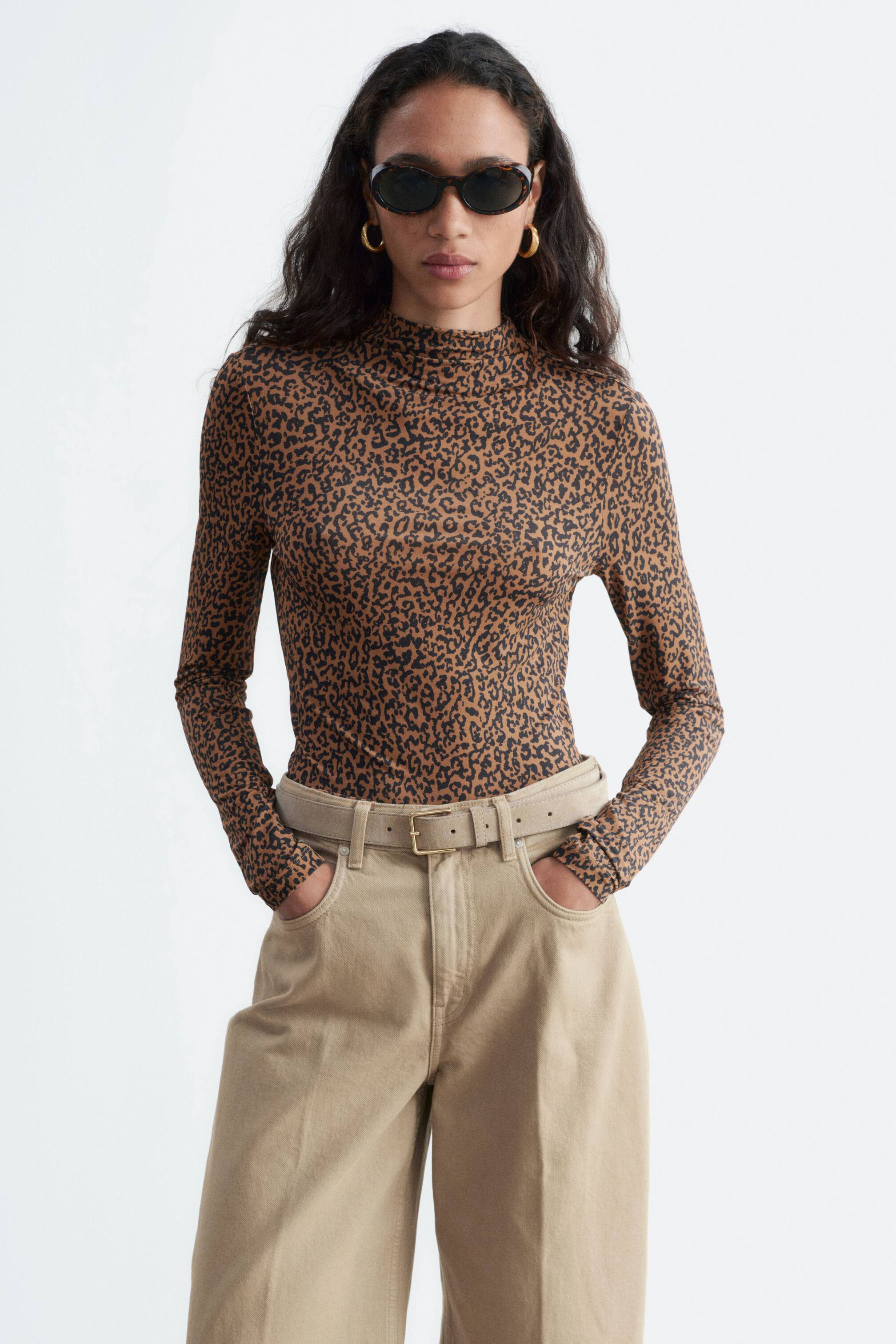 Agrandir l'image: Un haut imprimé à manches longues à col montant simulé, orné d’un motif léopard brun, est porté par le mannequin, mettant en valeur sa silhouette ajustée. Le haut est rentré dans un pantalon beige taille haute, maintenu par une ceinture.