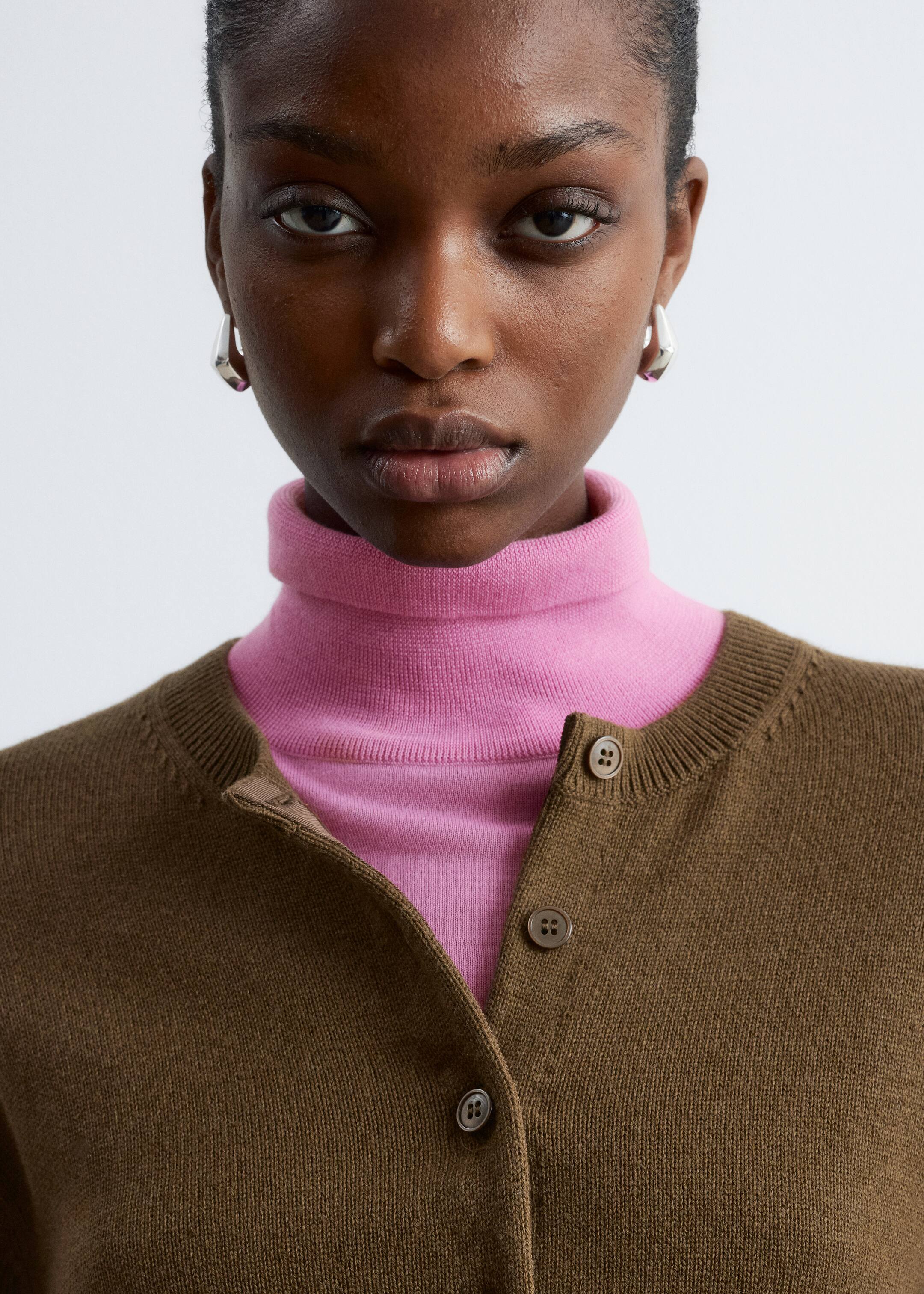 Se større billede: En brun crew-neck cardigan bæres over en lys pink turtleneck, med de to øverste knapper ubundne. Cardiganen har en fint strikket tekstur og synlige matchende brune knapper langs dens forplade.