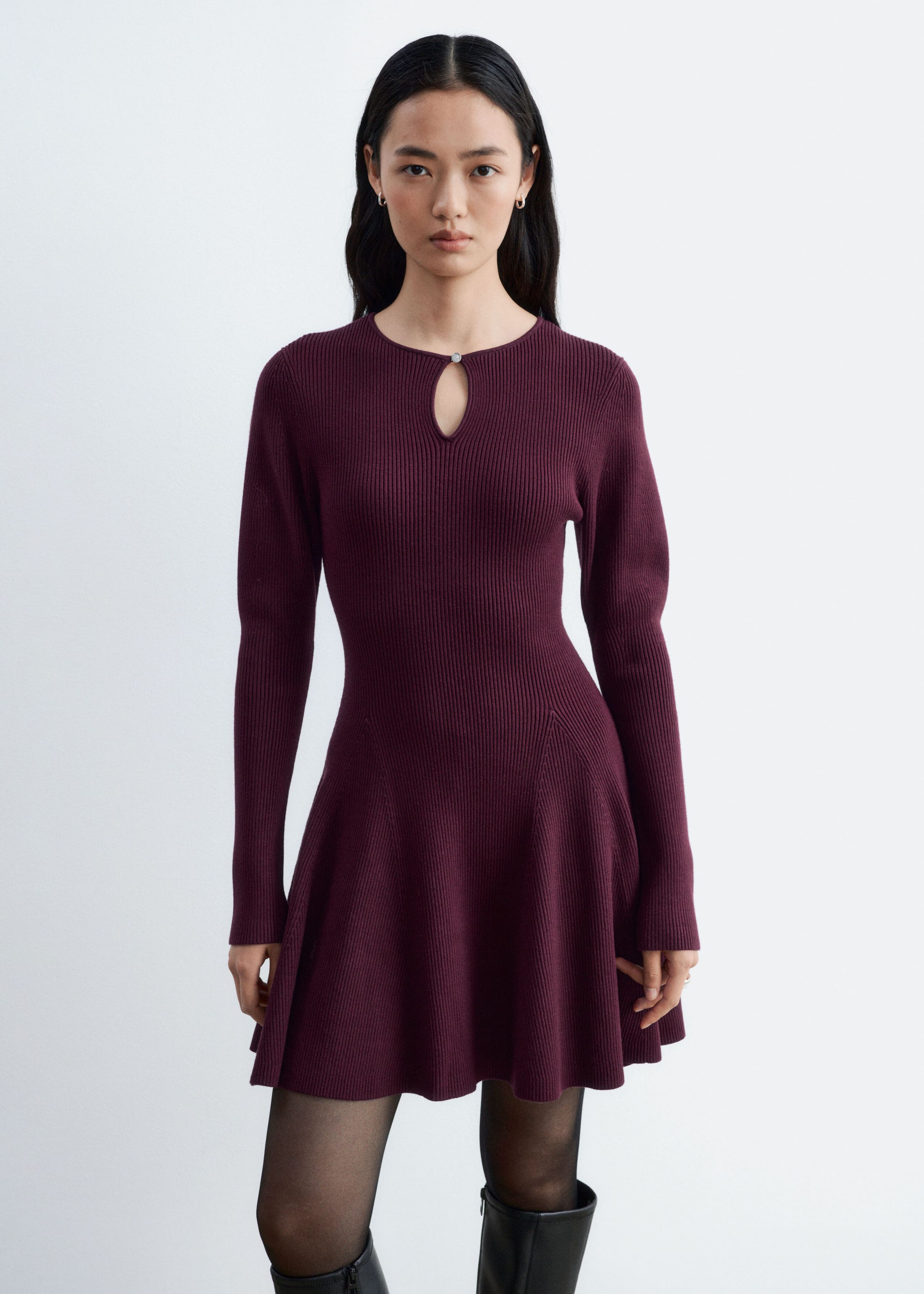 The model is wearing a plum-colored ribbed knit dress featuring long sleeves, a keyhole cutout at the neckline with a small metal button, and a flared paneled skirt. The dress is paired with sheer black tights and black knee-high boots.