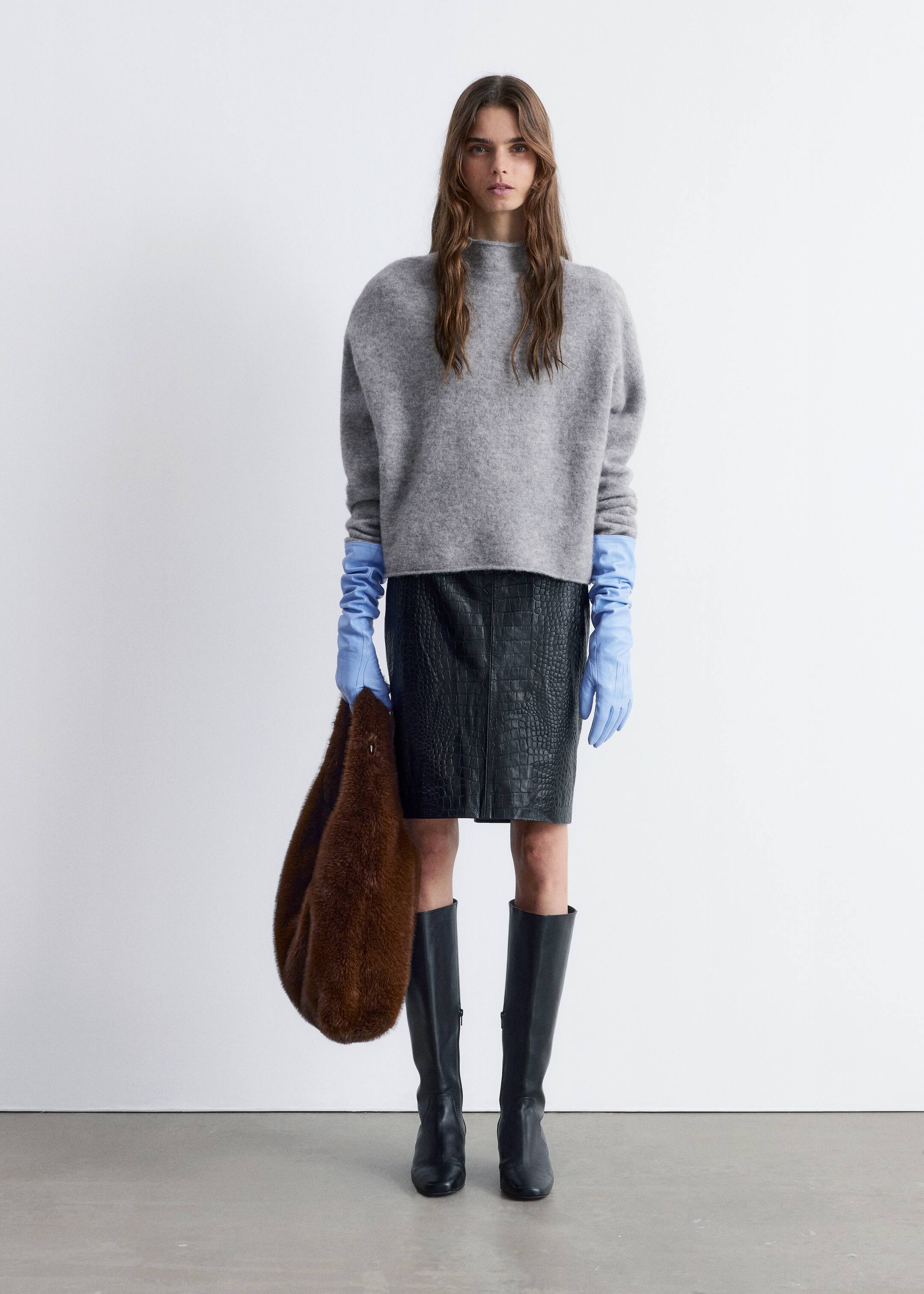 Se større billede: En grå, mock neck sweater bliver vist frem, med en afslappet, oversized pasform og sænkede skuldre. Dens lange ærmer strækker sig over lyseblå handsker og afslører en blød, fnugget tekstur.