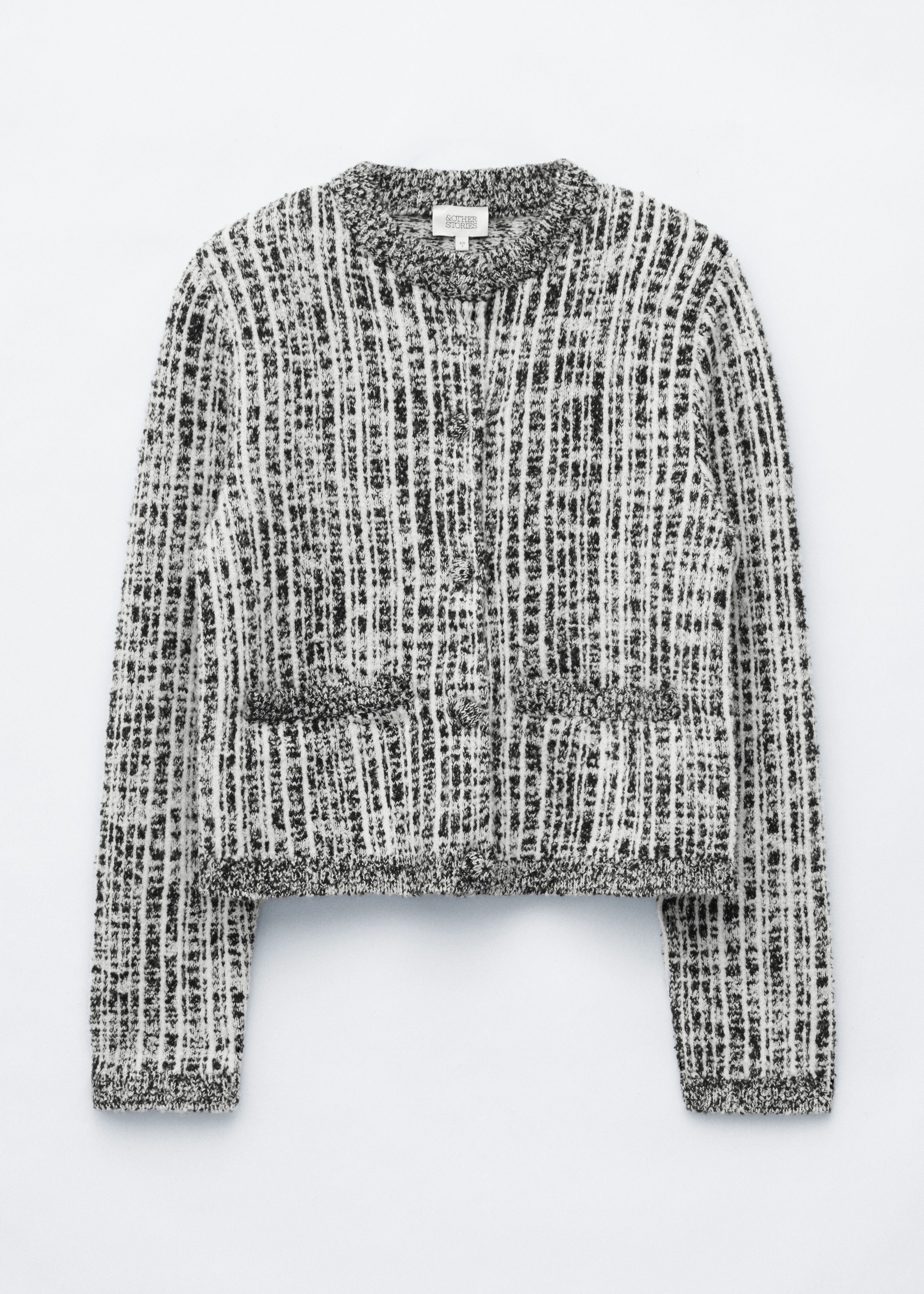A black and white textured knit cardigan featuring structured shoulders and ribbed trims is worn open by the model over a white T-shirt. The outfit is styled with dark grey jeans, and a red shoulder bag is carried under the model’s arm.