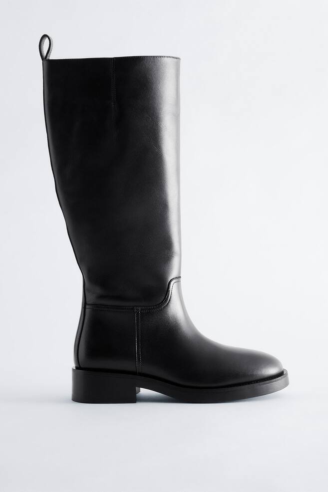 A knee-high black leather boot is shown in side profile, featuring a rounded toe, a low block heel, and a thick sole. The boot has a smooth leather finish with tonal stitching details around the ankle and a pull tab at the back of the collar. - 1