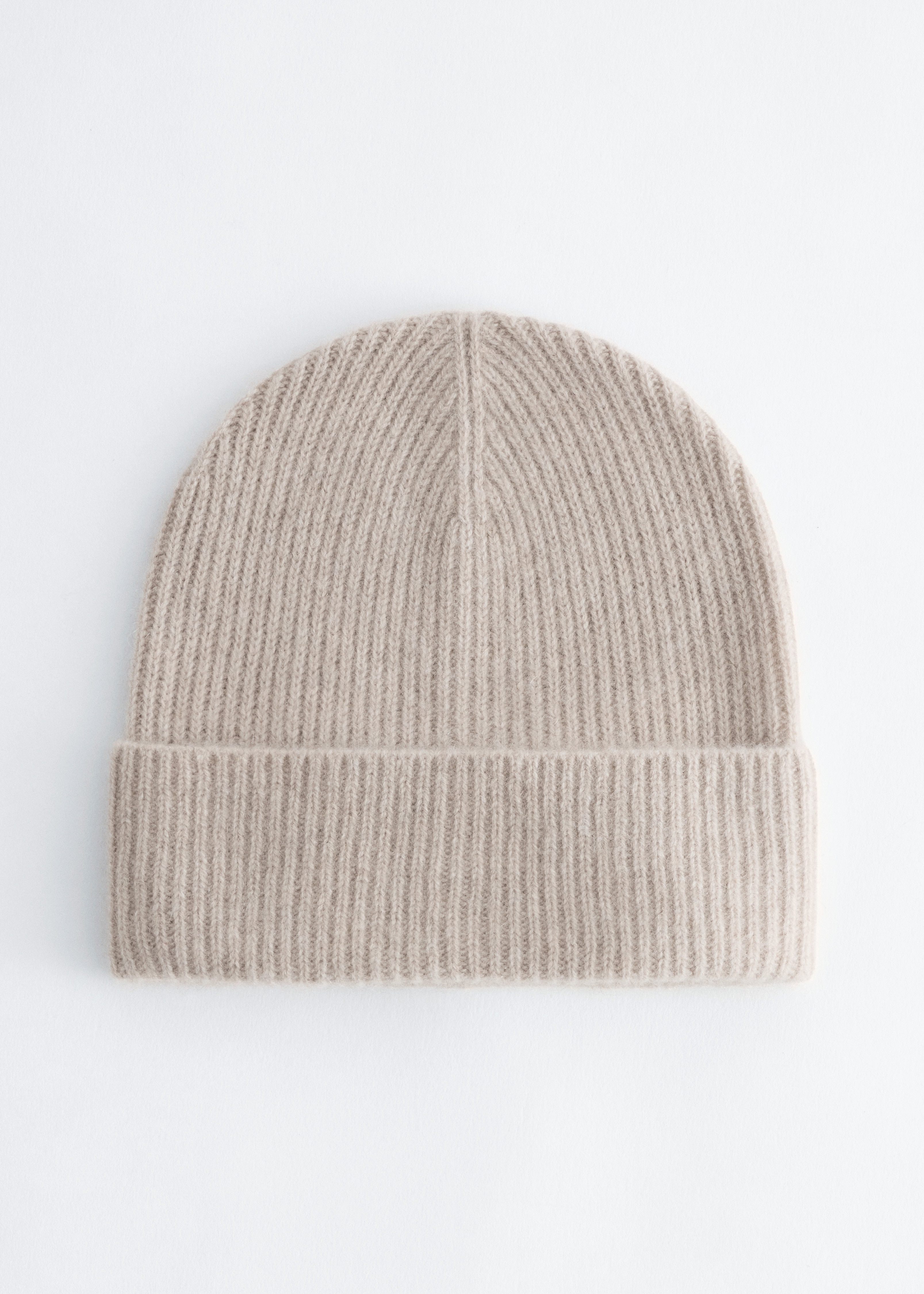 A light beige rib-knit beanie with a fold-over cuff is being worn on the modelâs head. The beanie is styled with a dark, textured coat collar visible at the bottom of the frame.