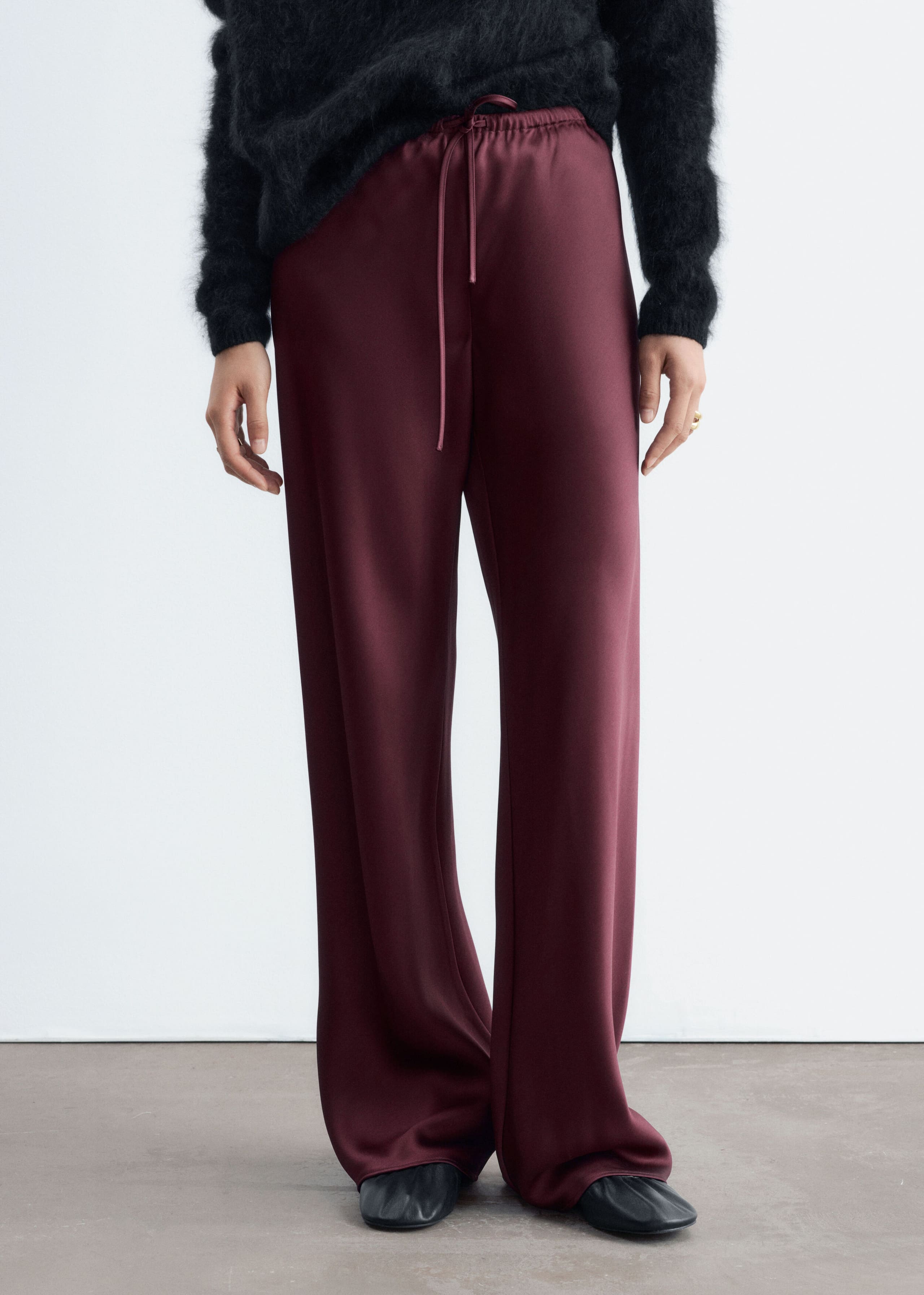 The model is wearing burgundy satin trousers with a relaxed, wide-leg silhouette, featuring an elastic waistband and a matching drawstring. The trousers are styled with a black fuzzy knit sweater tucked into the waist.