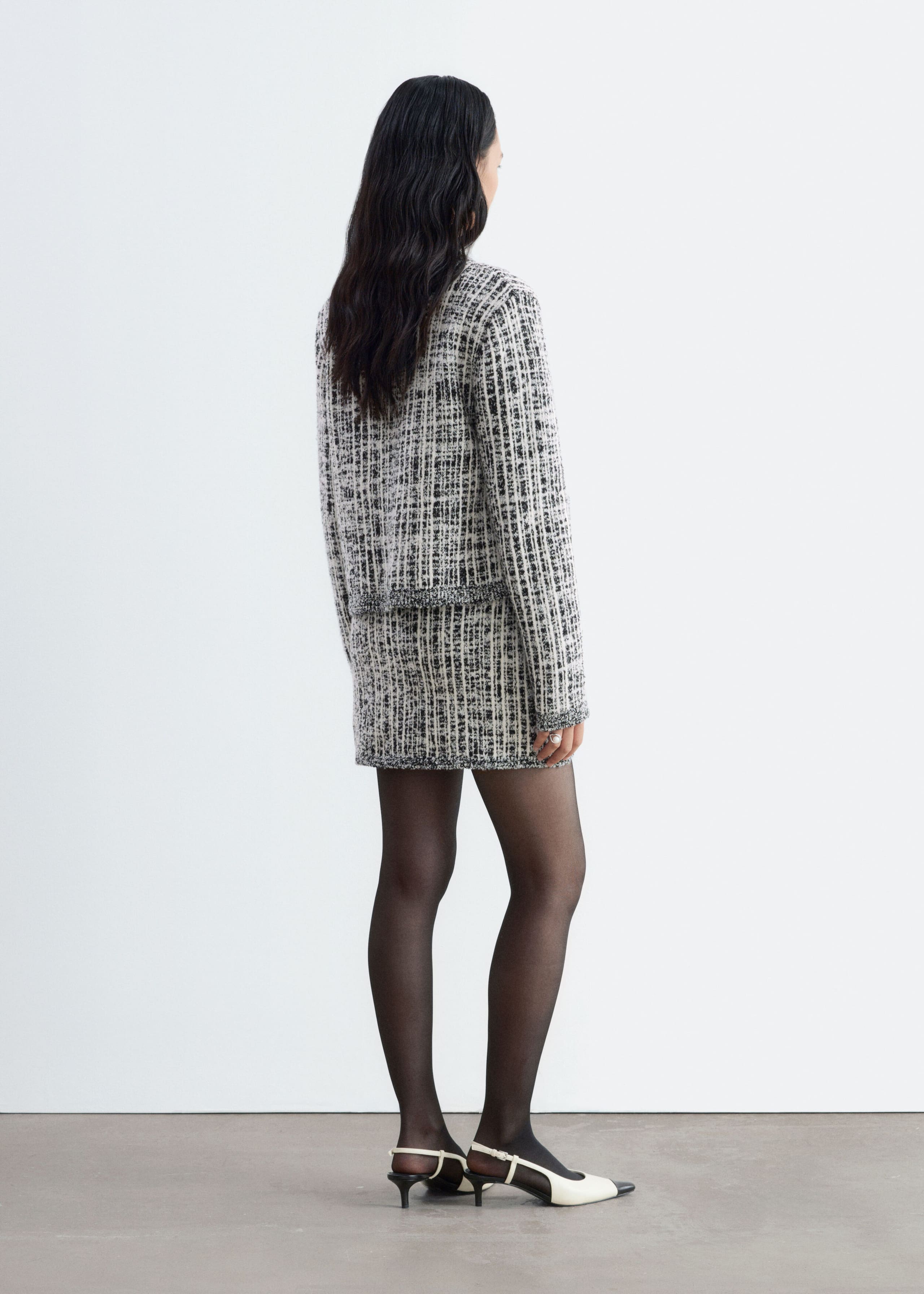 The model is wearing a black-and-white textured tweed jacket and matching miniskirt, shown from a three-quarter rear angle. The look is styled with sheer black tights and pointed-toe two-tone slingback heels.