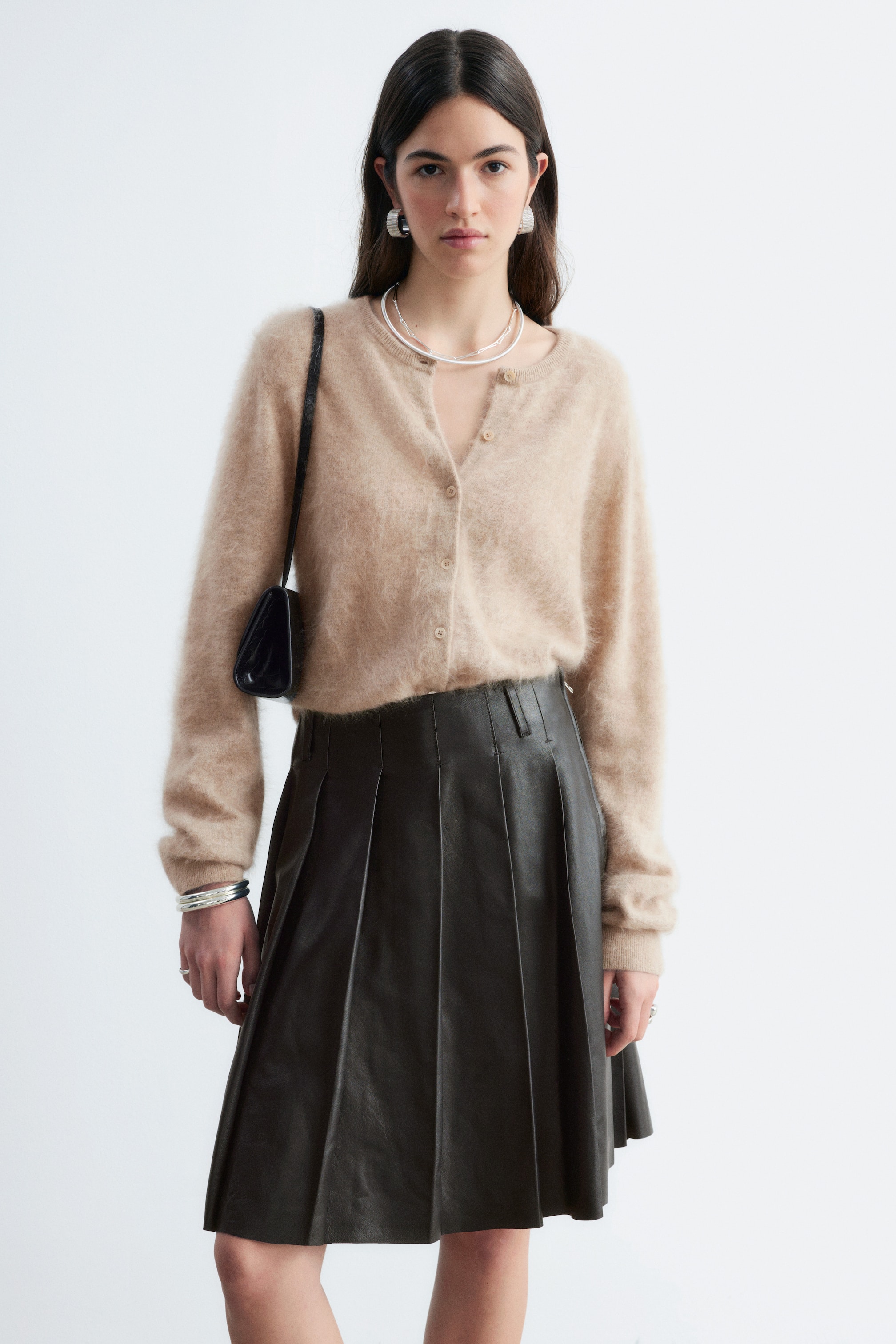 Grotere afbeelding bekijken: Een beige geborsteld kasjmier vest met een zachte, pluizige textuur wordt door het model dichtgeknoopt gedragen. Het vest is ingestopt in een donkere, geplooide leren rok en gestyled met een zwarte schoudertas.