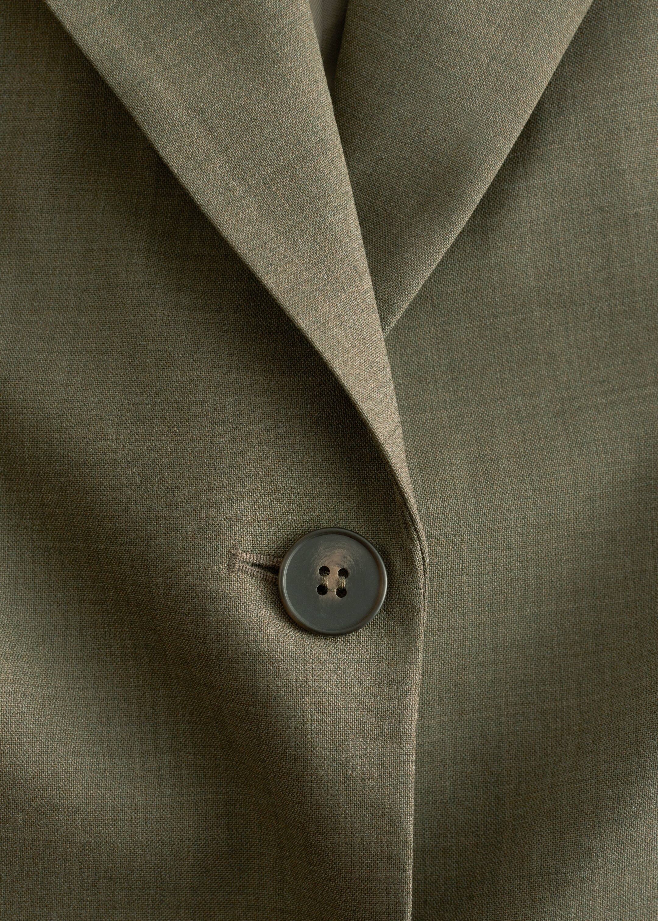 Visualizza immagine più grande: Una vista ravvicinata FRONTALE di un blazer monopetto in lana verde oliva mostra il suo risvolto con intaglio e un bottone scuro a quattro fori abbottonato.