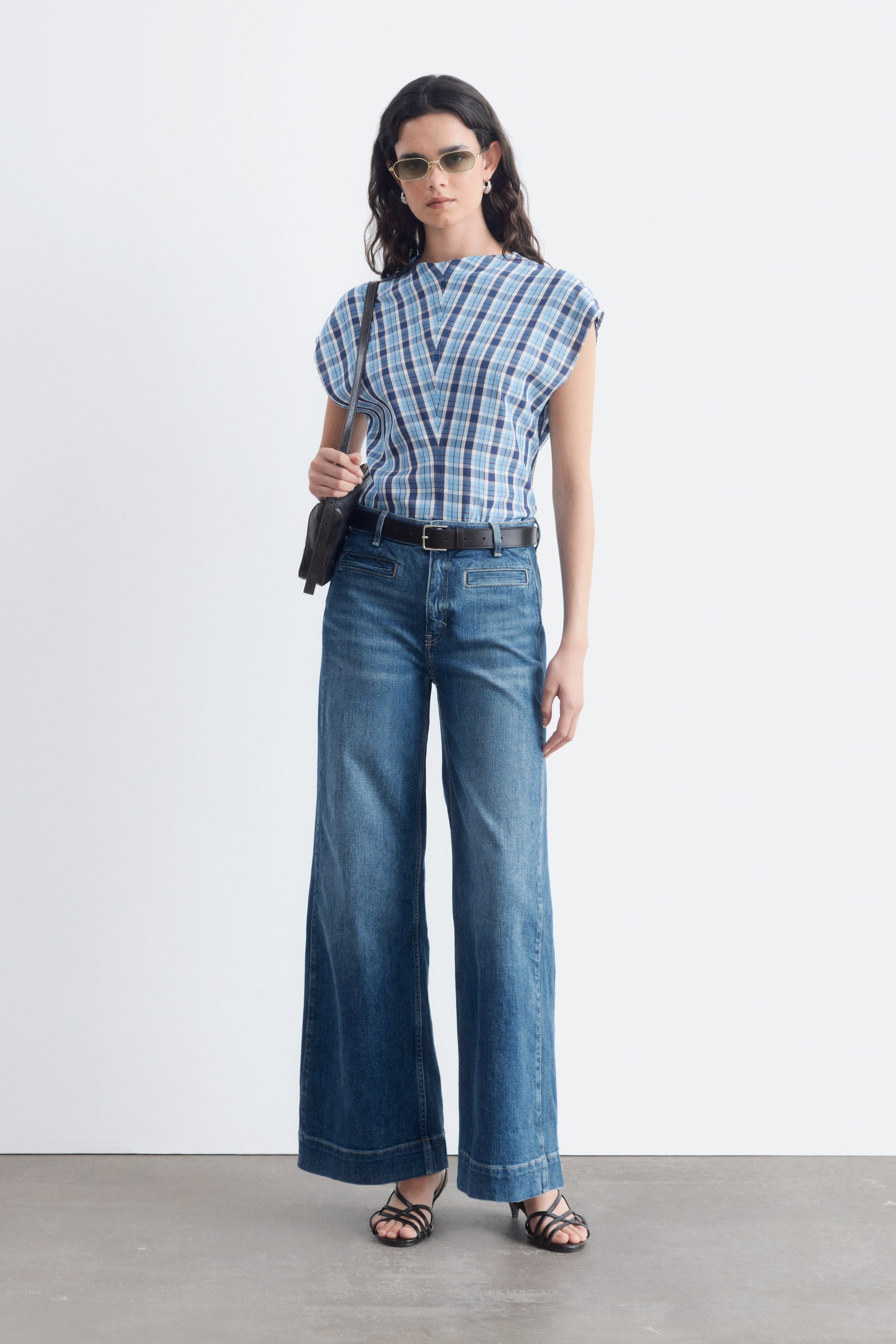 Agrandir l'image: Un jean wide-leg présente un délavage bleu moyen et une coupe taille haute, avec une silhouette ample et longue jusqu’aux chevilles. Il affiche un devant plat avec de subtiles surpiqûres, et une ceinture foncée est passée dans les passants de ceinture.