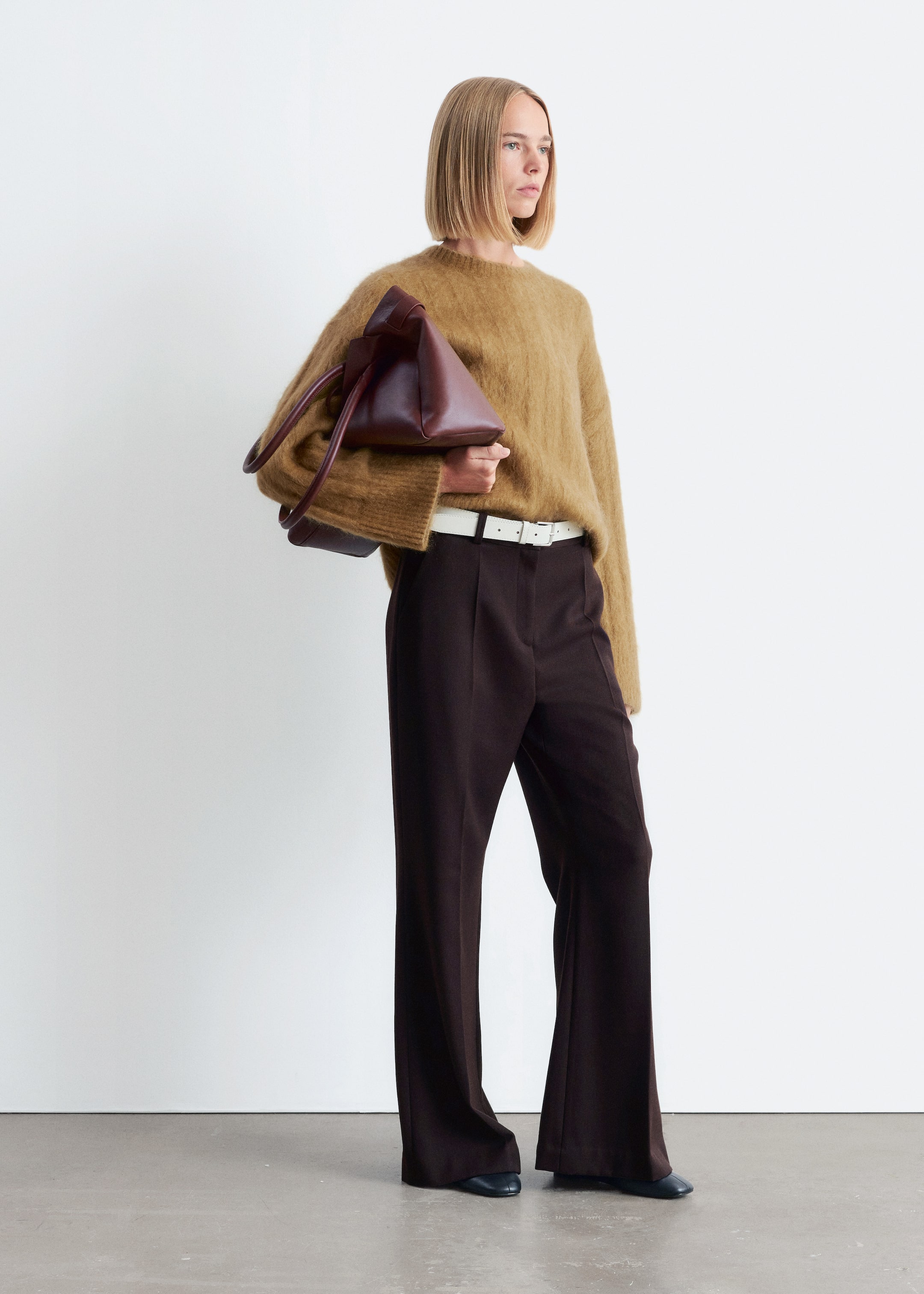 Agrandir l'image: Un pull ras du cou marron clair et duveteux est porté avec un pantalon large plissé marron foncé, qui est serré par une fine ceinture blanche. Un sac structuré brun rougeâtre foncé, avec deux poignées et une forme angulaire, est tenu, et des chaussures plates foncées sont visibles.