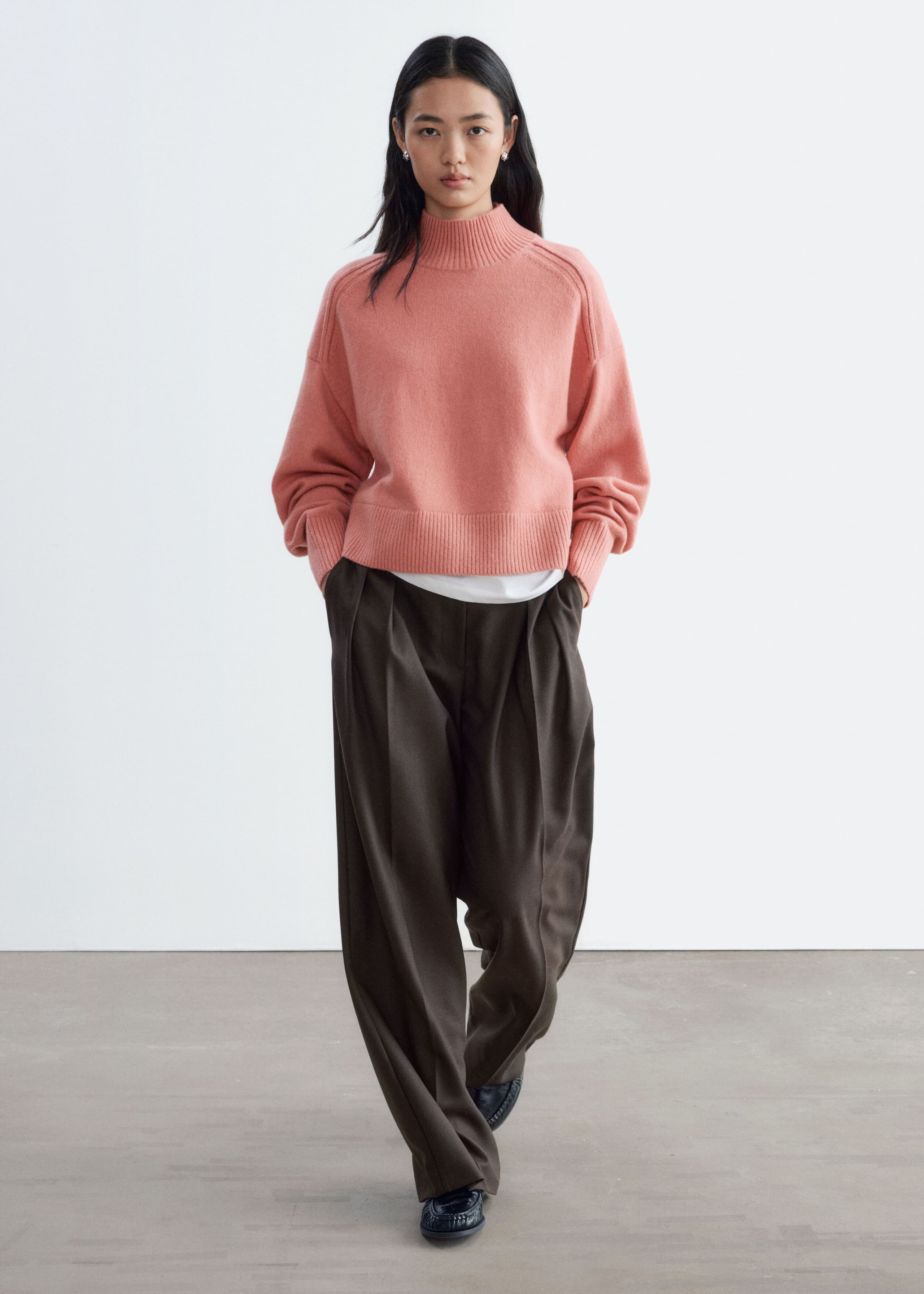 The model is wearing a pink mock neck sweater with a ribbed collar, hem, and cuffs. It is layered over a white T-shirt and paired with wide-leg, dark brown pleated trousers.