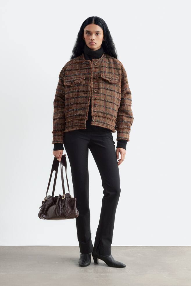 En brun tekstureret tweedjakke med vandrette striber, en kravelÃ¸s halsudskÃ¦ring, rÃ¥ kant-detaljer og brystlommer med klap bÃ¦res af modellen. Jakken er stylet over en sort rullekravesweater og sorte bukser, mens modellen holder en mÃ¸rkebrun lÃ¦derhÃ¥ndtaske. - 1