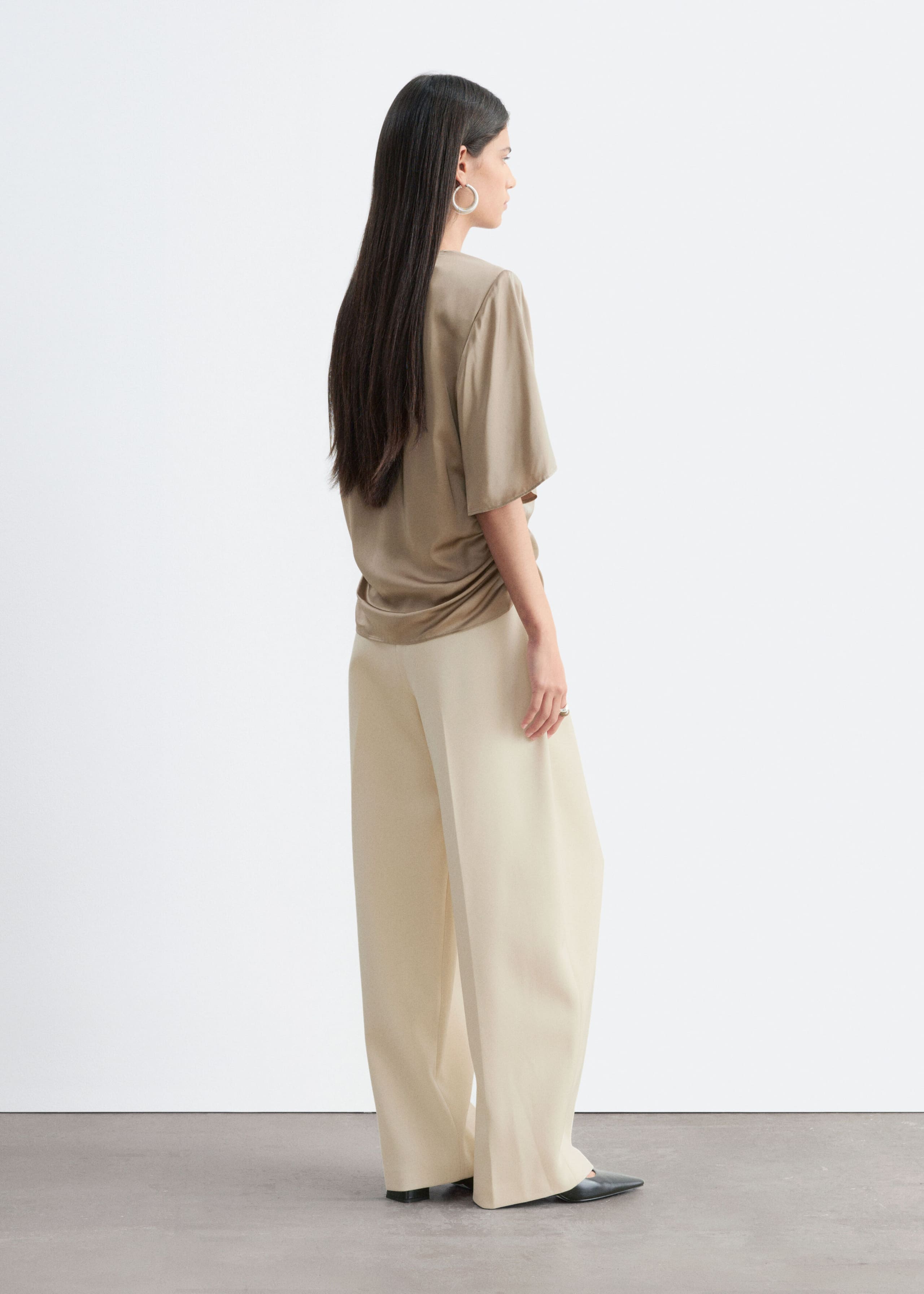 A taupe slim-fit blouse with soft shoulder pads and side pleats is worn by the model, who is standing in profile to show the back and drape of the garment. The blouse is styled with cream-colored wide-leg trousers.