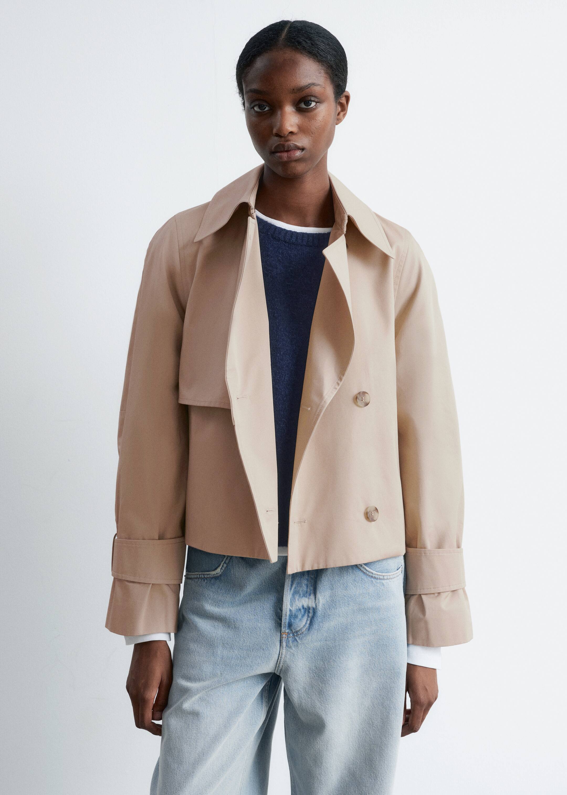 Ingrandisci l'immagine: Un trench coat corto beige è presentato frontalmente, indossato aperto sopra un maglione blu scuro e jeans a lavaggio chiaro. Presenta una chiusura doppiopetto con due bottoni visibili, ampi revers aperti, una pattina antipioggia sul petto sinistro e ampi cinturini ai polsi con fibbie che dettagliano le maniche.