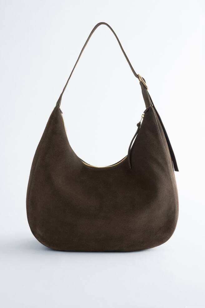 A dark brown suede shoulder bag is showcased from the front, boasting a rounded silhouette. It features a slim adjustable shoulder strap with a gold-toned buckle and fastens at the top with a gold-toned zip. - 1