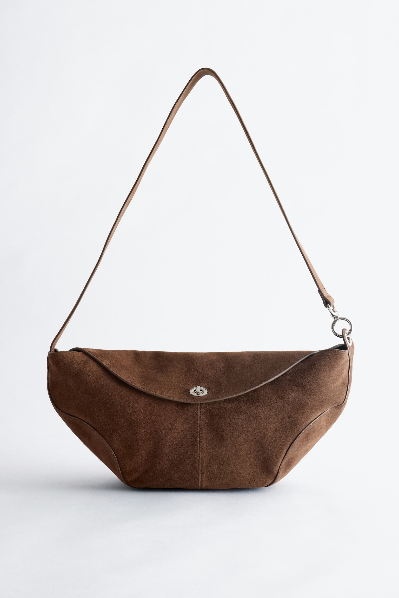 Na obrázku je zobrazená hnedá semišová crossbody kabelka z prednej strany, ktorá má sochársky tvar so zakriveným spodkom. Strieborný kovový otočný zámok zabezpečuje klapku v štýle obálky a tenký hnedý popruh je pripevnený na každej strane. - 1