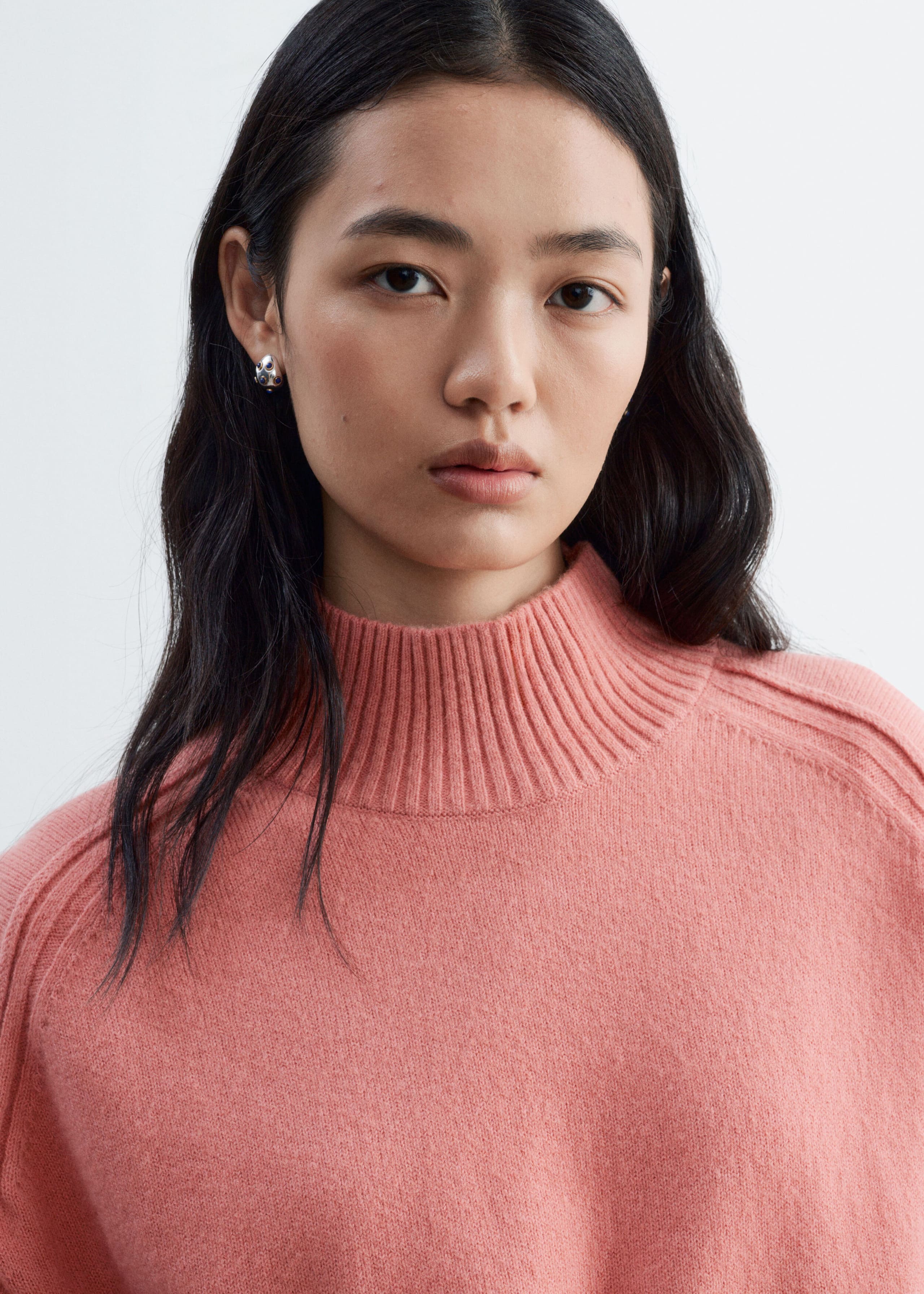 A pink knit sweater is being worn by the model, featuring a high ribbed mock neck and forward-set shoulder seams. The soft, textured fabric is shown in a close-up view.