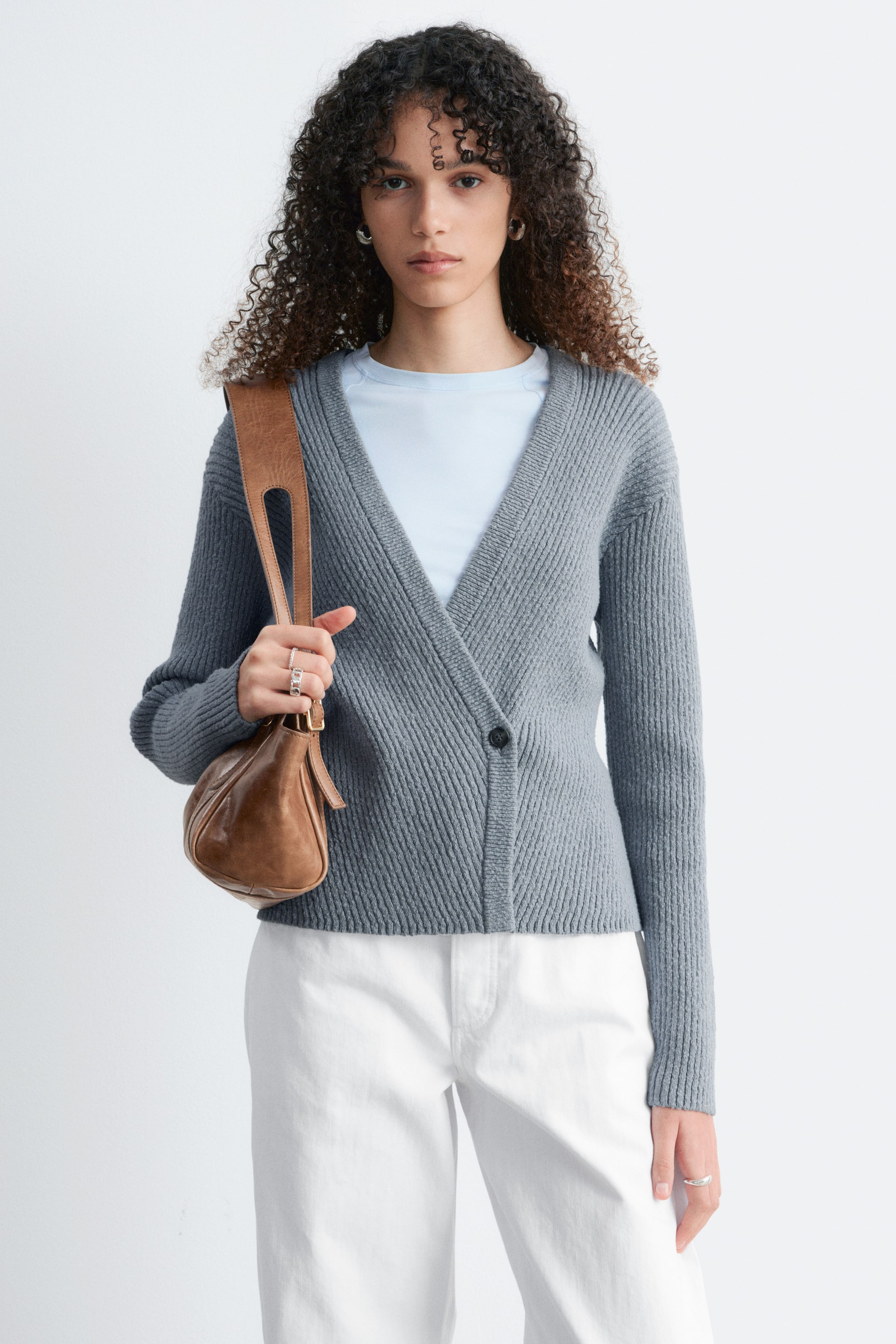 Nagyobb kép megtekintése: A szürke bordázott kötött kardigán V-nyakkivágással és aszimmetrikus átlapolt stílussal rendelkezik, amelyet egyetlen fekete gomb rögzít, és egy világoskék póló fölött viselik.