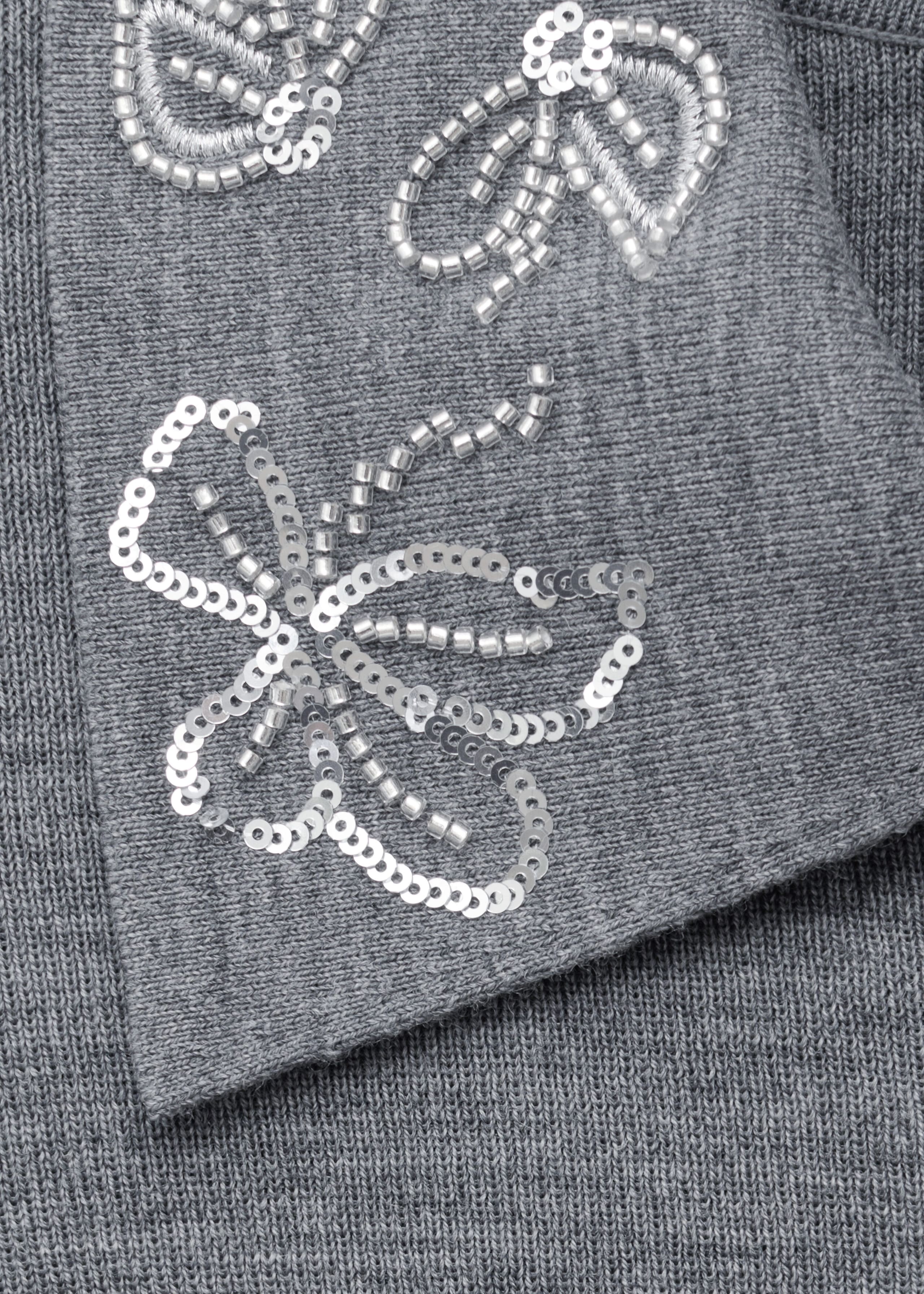 Eine Nahaufnahme eines grauen Woll-Polo-Pullovers zeigt das Kragendetail, das mit silbernen Pailletten und zylindrischen Perlen in floralen Mustern verziert ist. Der fein gestrickte graue Stoff bringt seine weiche Struktur zur Geltung, während der Kragen über das Kleidungsstück umgeschlagen liegt.