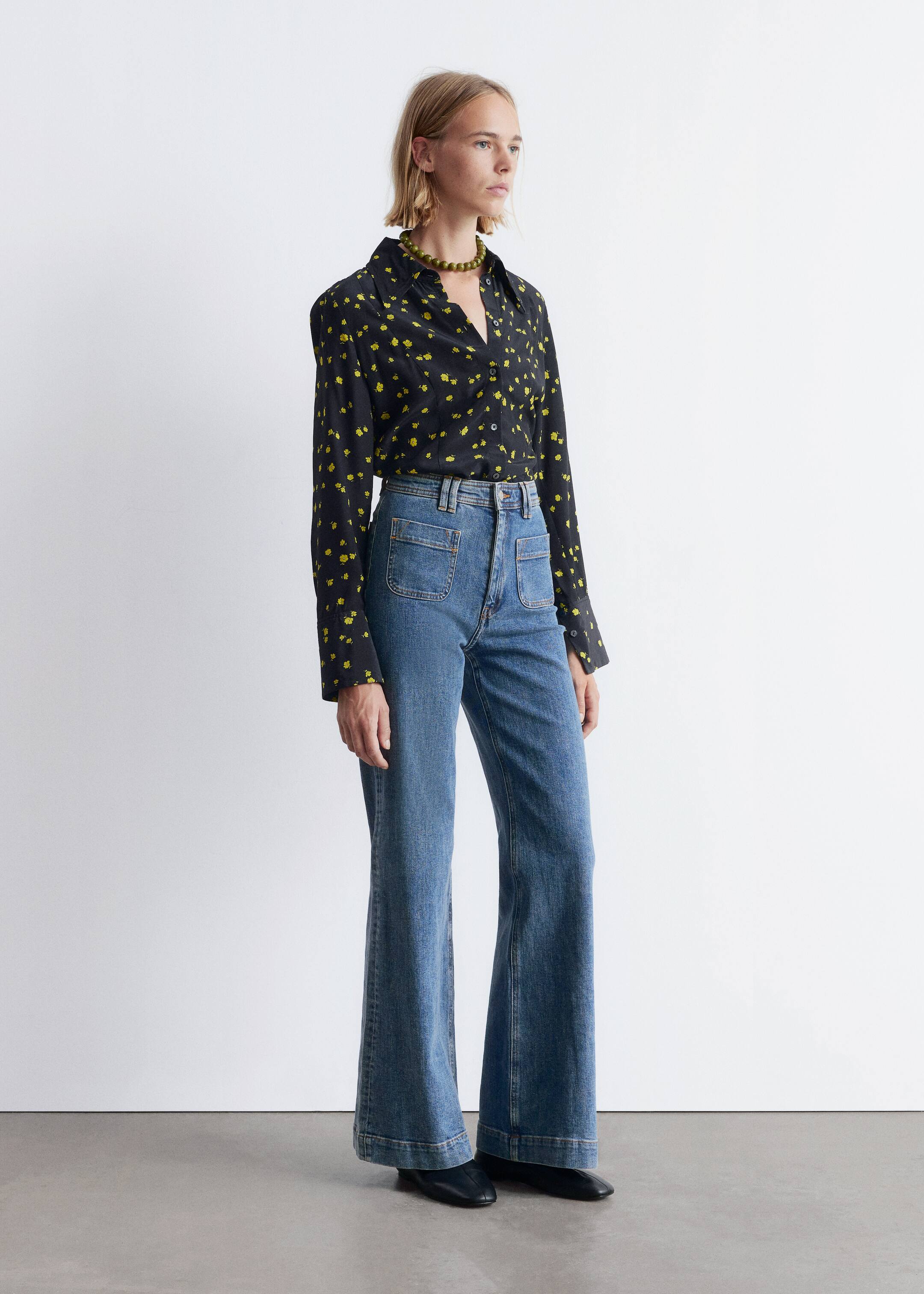 Grotere afbeelding bekijken: Een zwarte overhemd met knopen en een gele bloemenprint wordt gepresenteerd in een driekwartaanzicht, met een puntige kraag en lange mouwen. Het wordt gestyled met blauwe hoog getailleerde flared jeans, die grote voorzakken hebben en aanzienlijk uitlopen naar de zoom, waardoor zwarte platte schoenen bedekt worden.