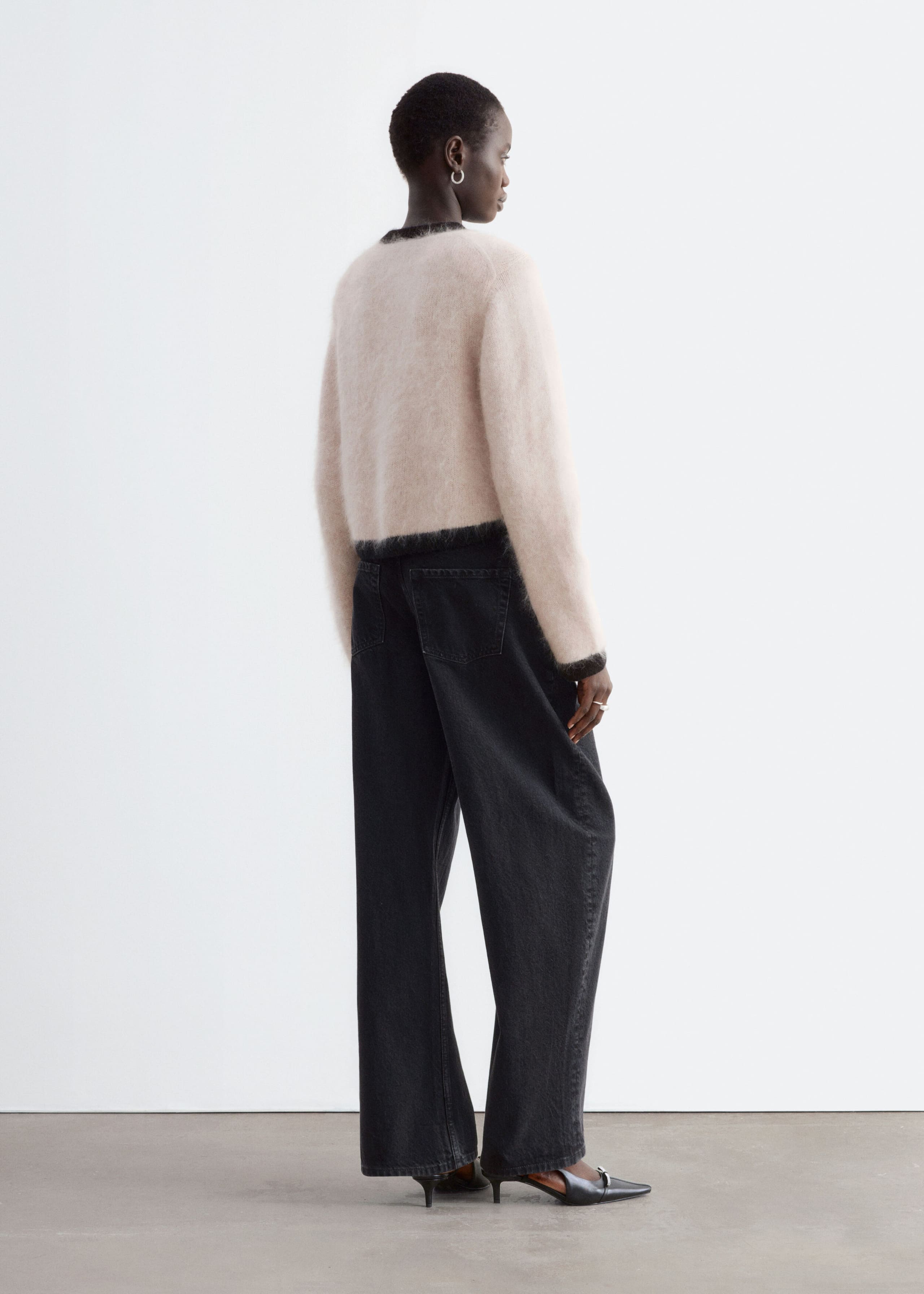 A fuzzy, light pink knit cardigan with black ribbed trim along the collar, hem, and cuffs is worn by the model. The cardigan features a structured shoulder silhouette and is styled with high-waisted, wide-leg black jeans.