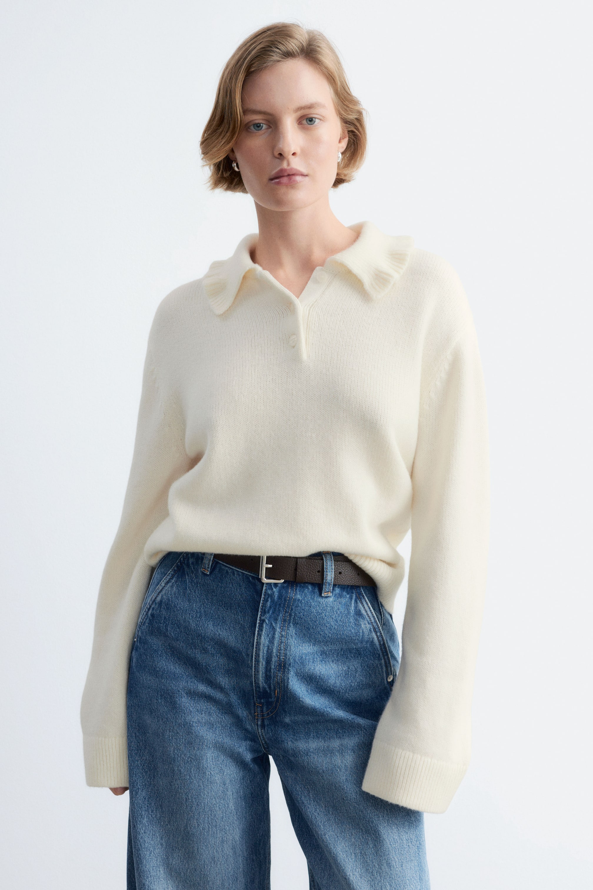 Größeres Bild ansehen: Ein cremefarbener Strickpullover mit gerüschtem Polokragen und Knopfleiste wird vom Model getragen. Das langärmlige Oberteil ist in blaue Jeans gesteckt, die mit einem braunen Gürtel kombiniert sind.