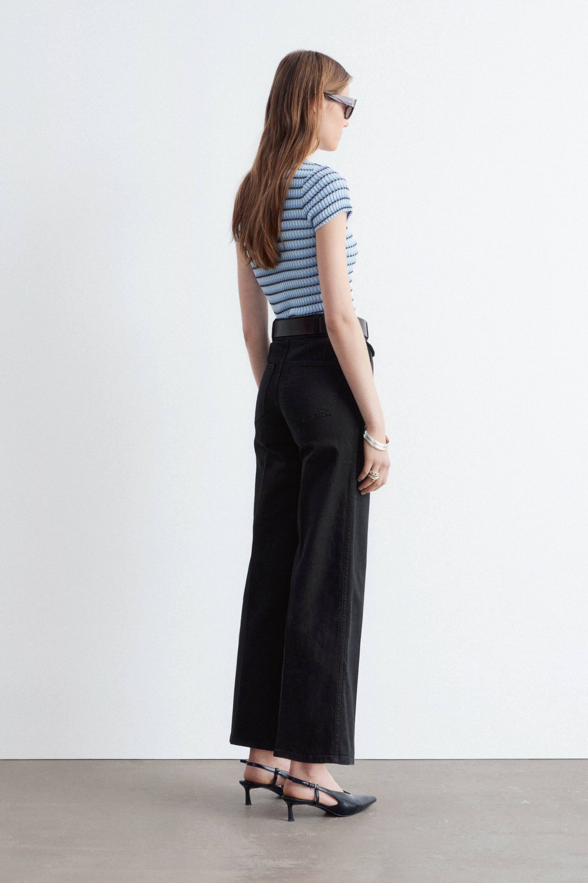 Größeres Bild ansehen: Die schwarze High-Waist-Jeans mit ausgestelltem Bein, Bügelfalten und zwei aufgesetzten Gesäßtaschen wird vom Model getragen. Ein Gürtel ist durch die sichtbaren Gürtelschlaufen der Jeans gezogen, und die Jeans ist in voller Länge.