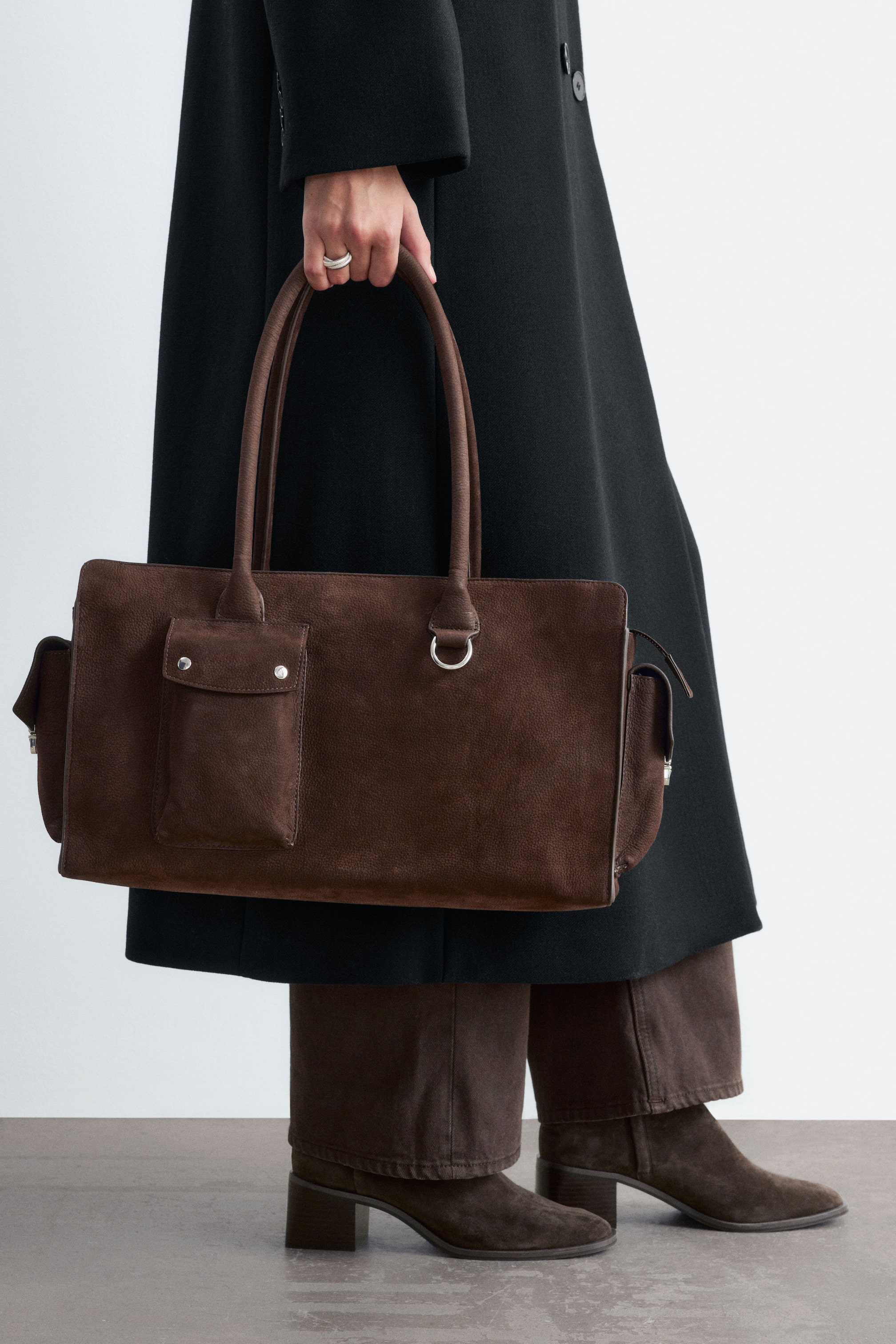 Grotere afbeelding bekijken: Een bruine rechthoekige totebag in suède wordt aan de bovenste handvatten vastgehouden door het model, dat een lange zwarte jas, bruine broek en bijpassende suède laarzen draagt. De tas heeft aan de voorkant een envelopzak met twee zilveren drukknoopsluitingen, een metalen D-ringdetail en gestructureerde zijpanelen.