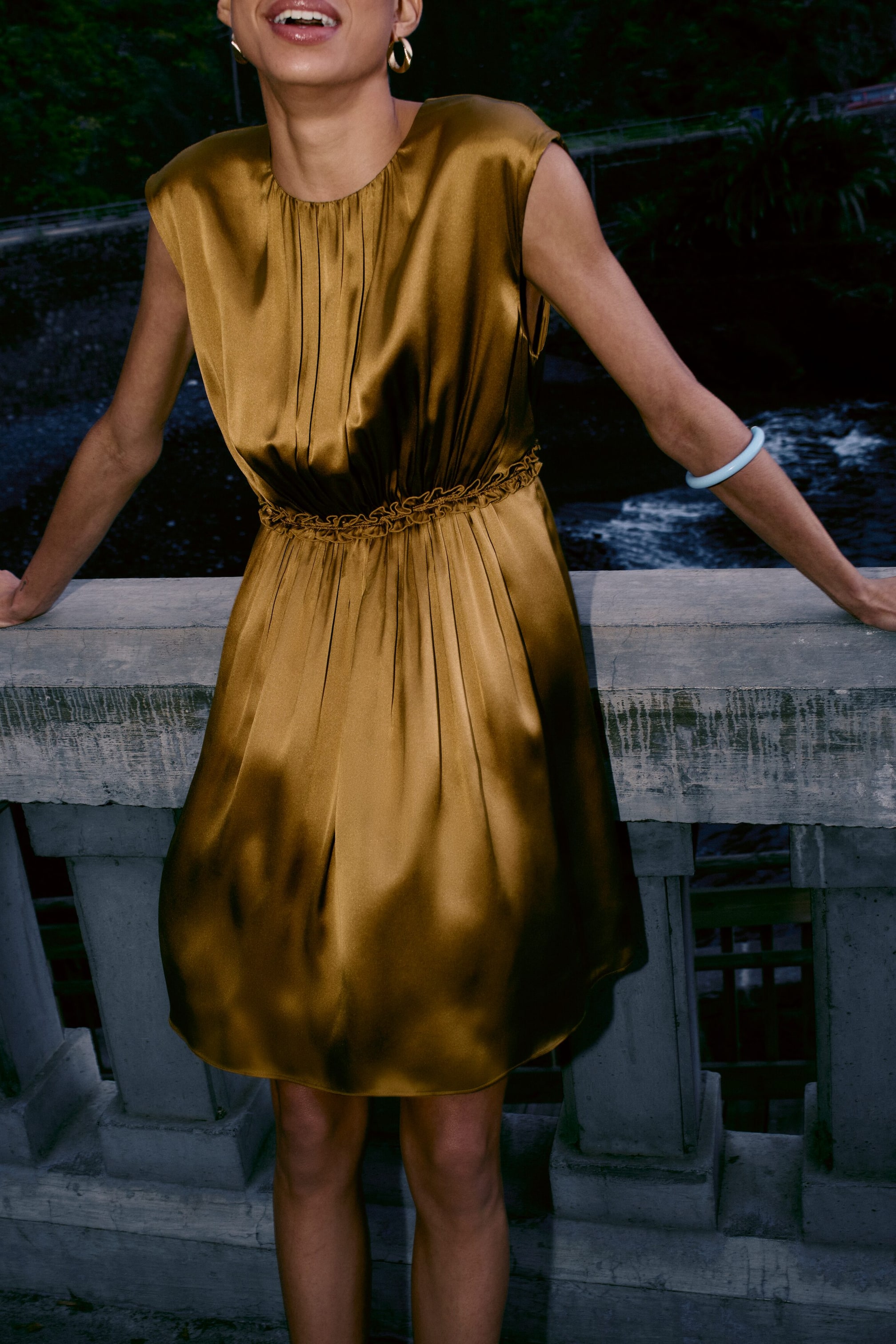 Ver imagen más grande: Un vestido de satén en color bronce dorado presenta un escote redondo fruncido y mangas tipo capa. El vestido se ajusta en la cintura con un detalle fruncido y fluye en una falda de línea A, que termina por encima de la rodilla.