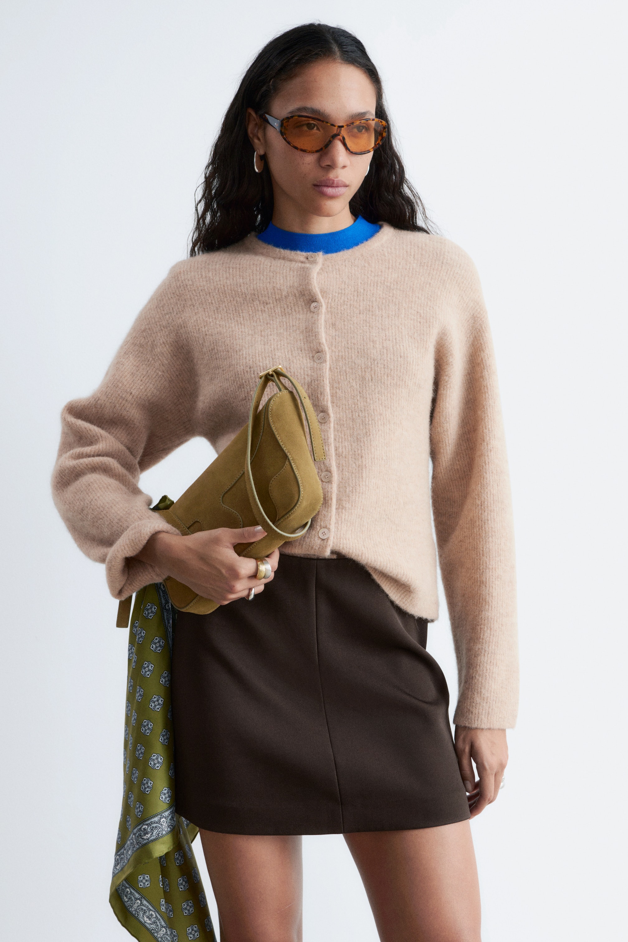 Преглед на по-голямо изображение: Бежовият кардиган се отличава с пухкава плетена текстура и кръгло деколте, като се закопчава отпред с малки, светли копчета. Представя се с отпусната кройка и дълги ръкави, а в областта на деколтето се вижда син топ.