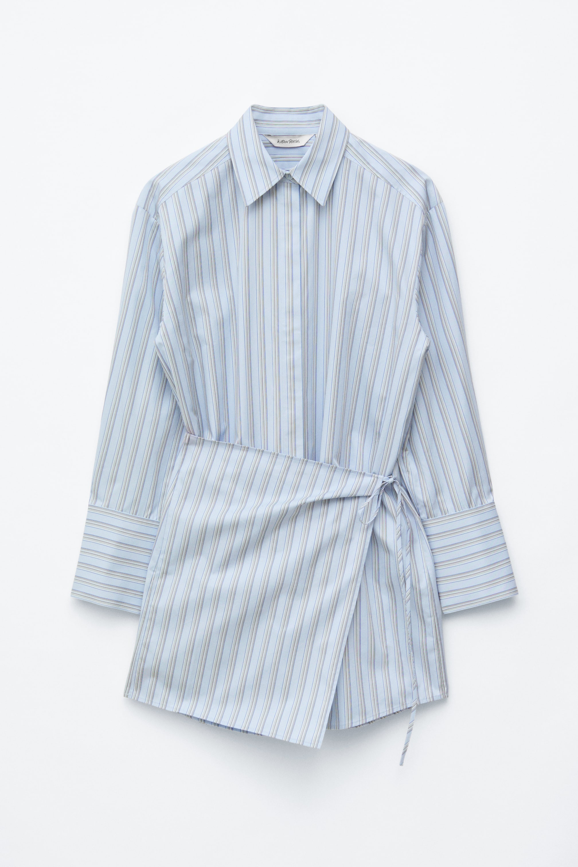 Ver imagen más grande: Se muestra desde el frente un vestido camisero mini de color azul claro con rayas verticales. Presenta un cuello, una tapeta con botones, mangas largas con puños abotonados y una cintura asimétrica con cierre de lazo en el lado izquierdo.