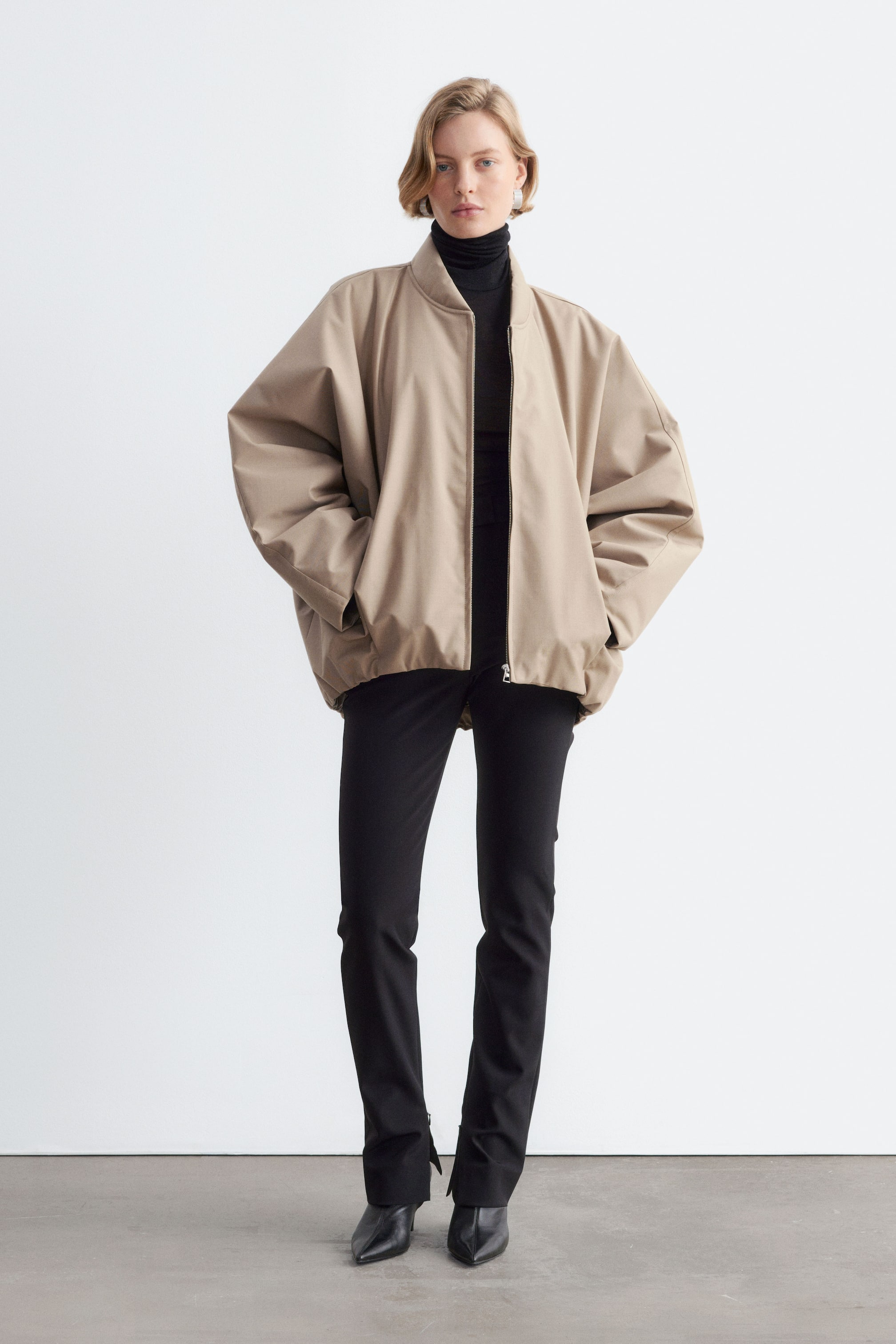 Größeres Bild anzeigen: Ein oversized beiger Bomberjacke mit voluminösen Ärmeln und elastischem Saum wird vom Model getragen, das mit den Händen in den Seitentaschen steht. Die Jacke ist über einem schwarzen Rollkragenoberteil und einer eng anliegenden schwarzen Hose kombiniert.
