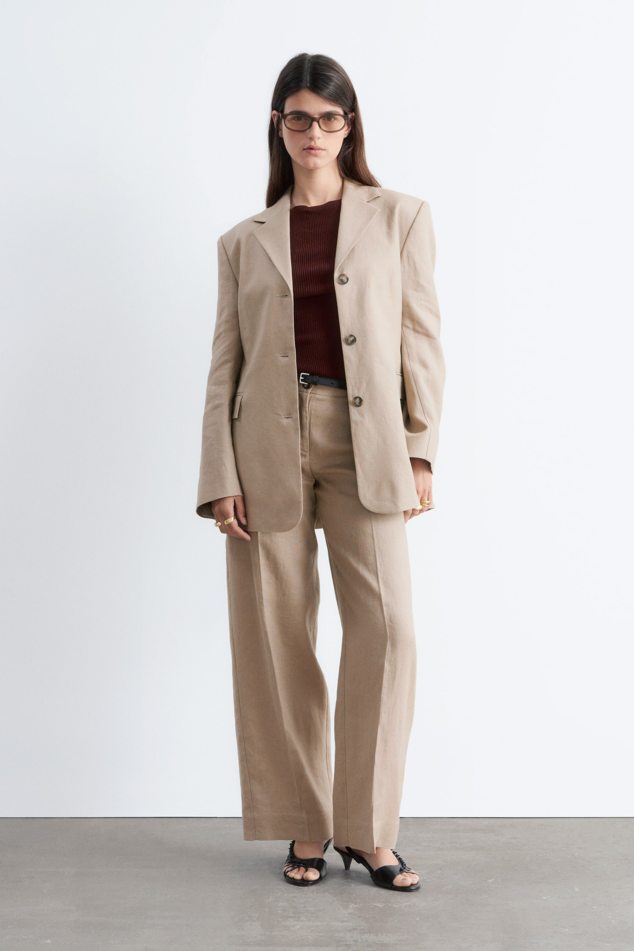 Agrandir l'image: Un costume en mélange de lin beige est présenté, comprenant un blazer à simple boutonnage avec des revers crantés, trois boutons, des épaules structurées et deux poches plaquées. Le pantalon assorti à jambes larges complète le costume, affichant un pli tailleur et étant porté avec une ceinture foncée sur un haut côtelé rouge foncé.