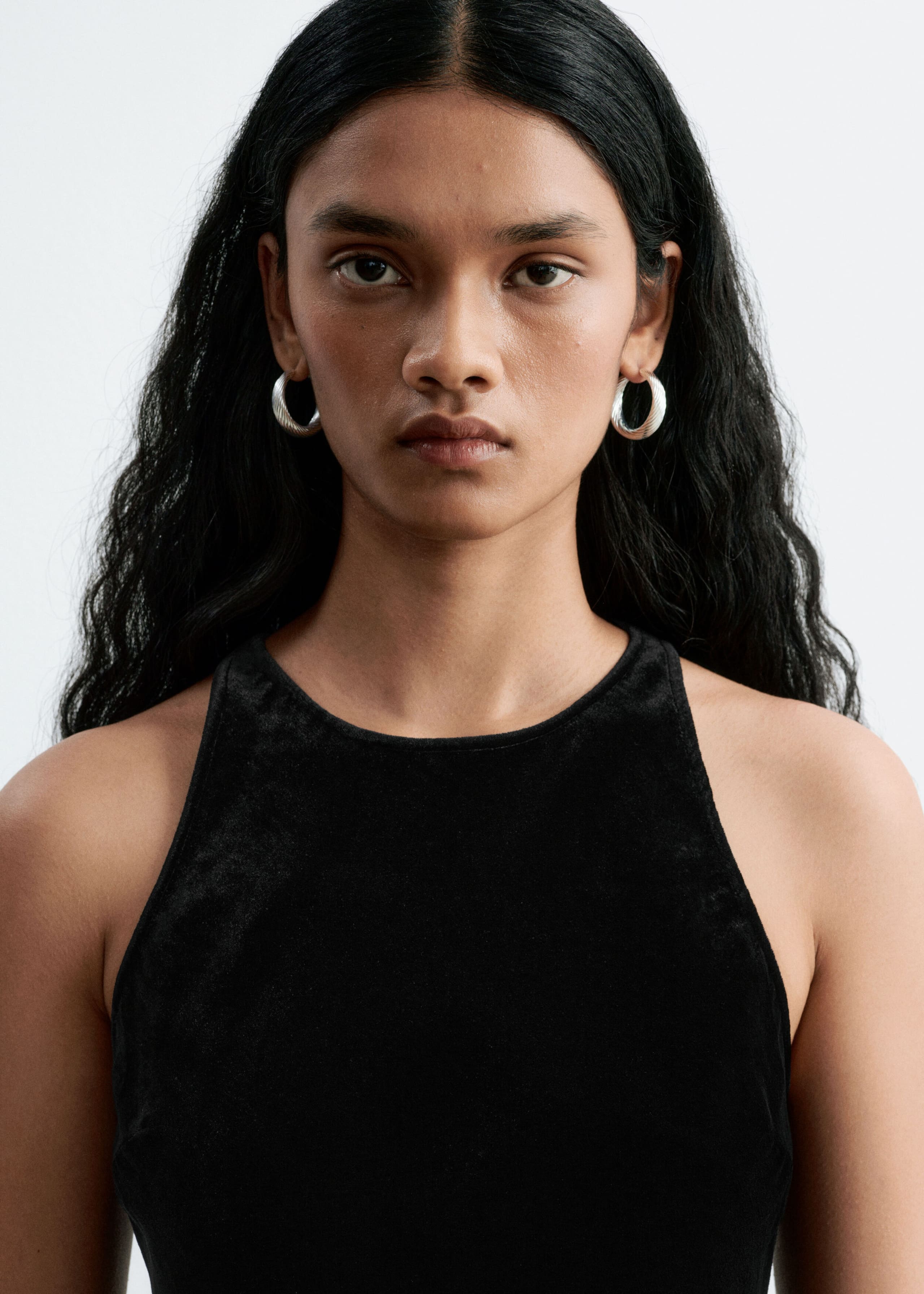 A black sleeveless dress with a high crew neckline is being worn by the model in a close-up view. The dress features a soft, textured finish and is styled with silver hoop earrings.
