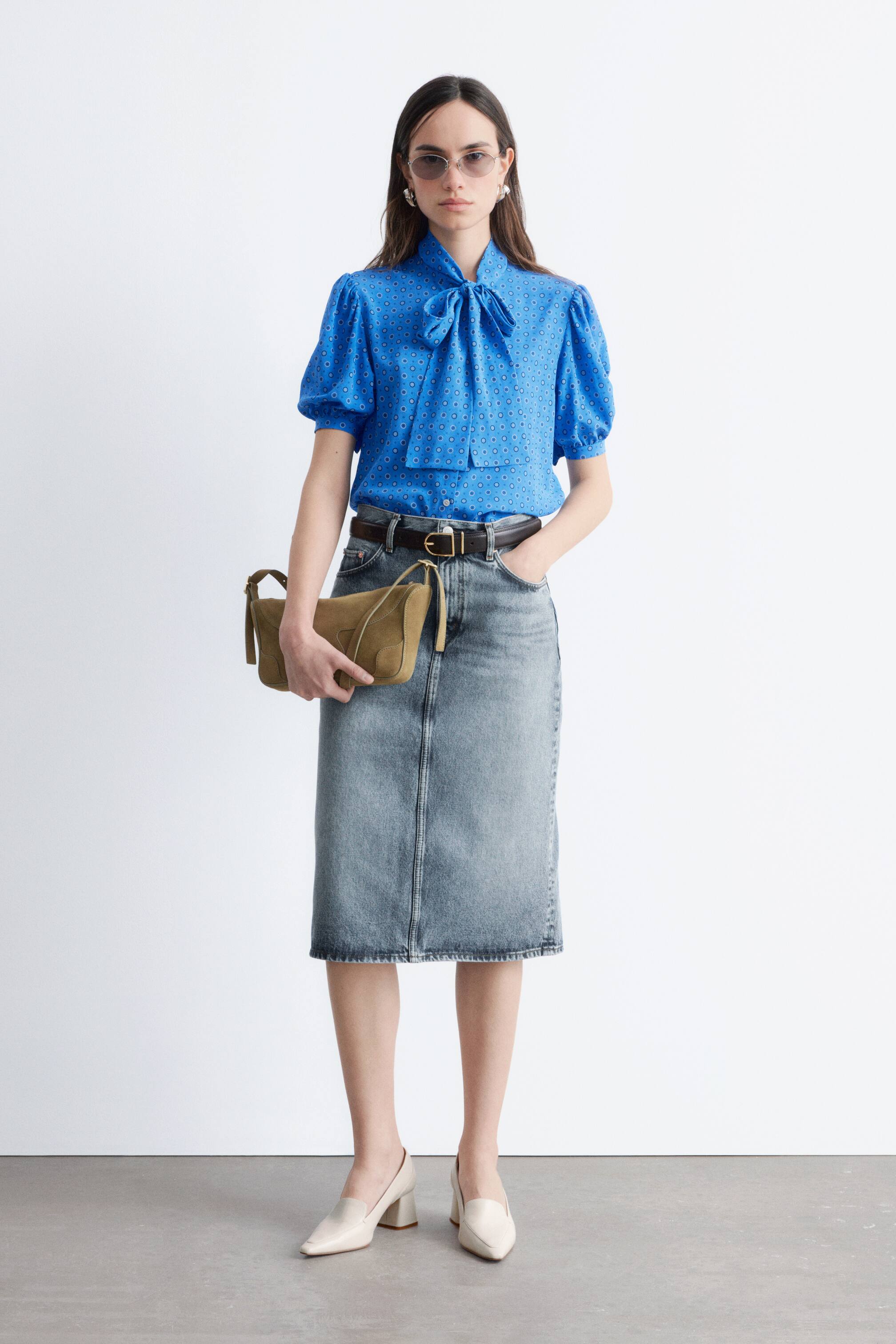 Visualizza immagine più grande: Una blusa blu presenta un piccolo motivo geometrico circolare, un colletto a fiocco e maniche corte a sbuffo. È frontale e infilata in una gonna di jeans.