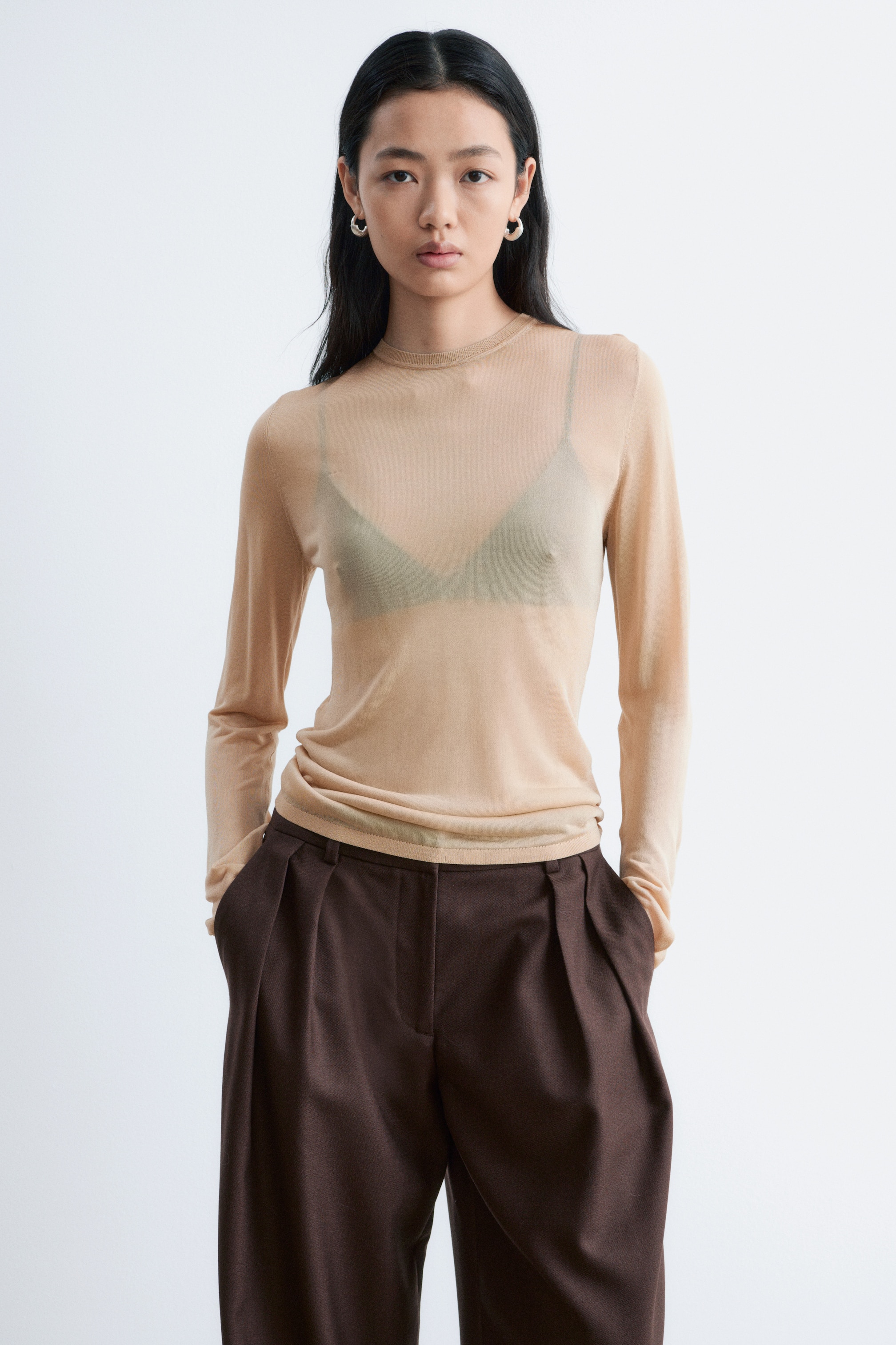 Agrandir l'image: Un haut transparent à manches longues de couleur beige clair, avec un col rond et des finitions côtelées, est porté par le mannequin. Un soutien-gorge foncé est visible sous le haut, qui est associé à un pantalon brun foncé.