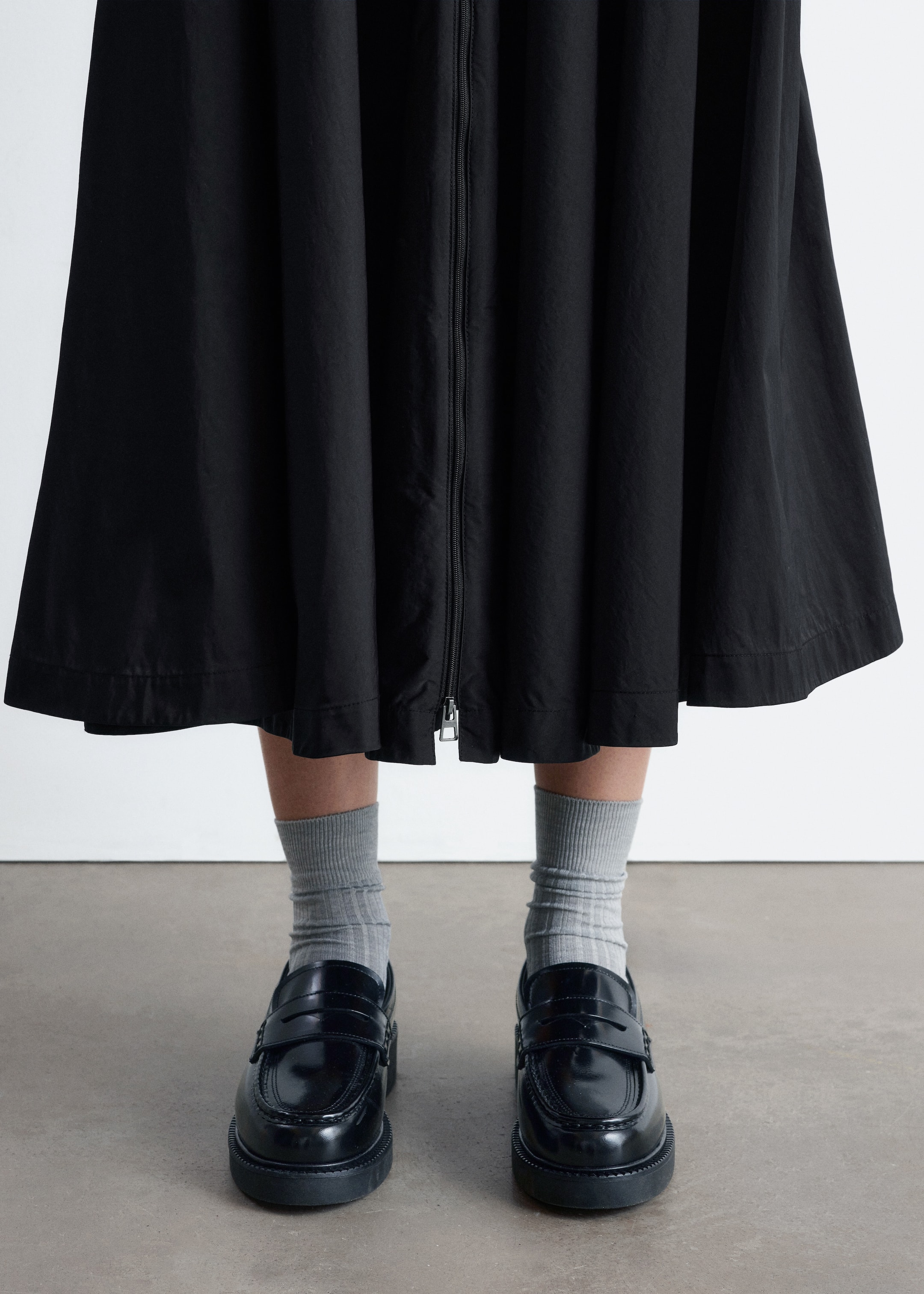 Agrandir l'image: Des mocassins en cuir verni noir avec des semelles épaisses sont portés, orientés vers l'avant, associés à des chaussettes côtelées gris clair. Un vêtement noir, possiblement une robe ou une jupe, est suspendu avec une coupe ample, doté d'une fermeture éclair à l'avant qui est ouverte de l'ourlet vers le haut.