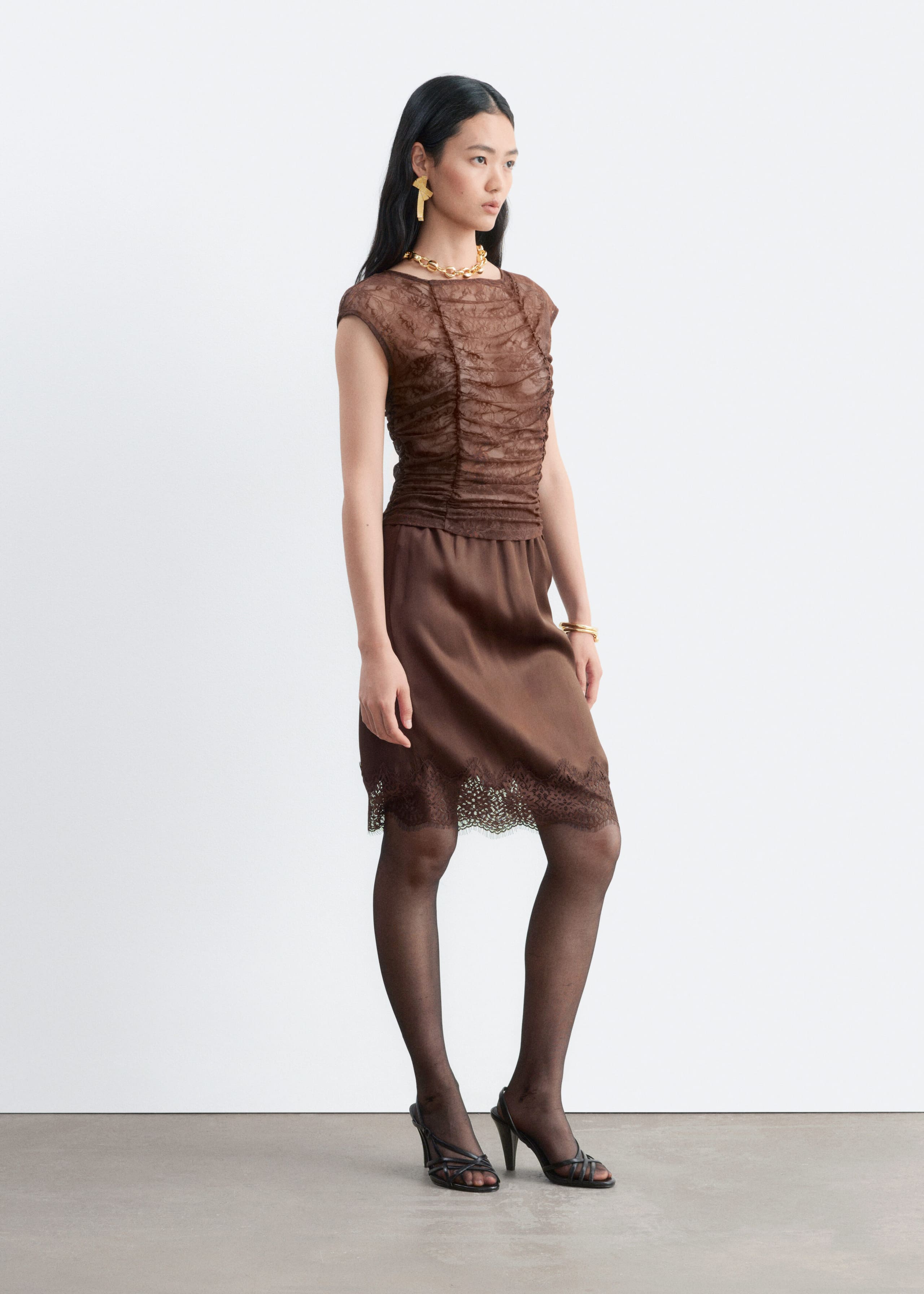 A brown lace top featuring ruching through the body, cap sleeves and a boat neckline is being worn by the model. The top is styled with a matching brown satin skirt with lace trim, black sheer tights and black heeled sandals.