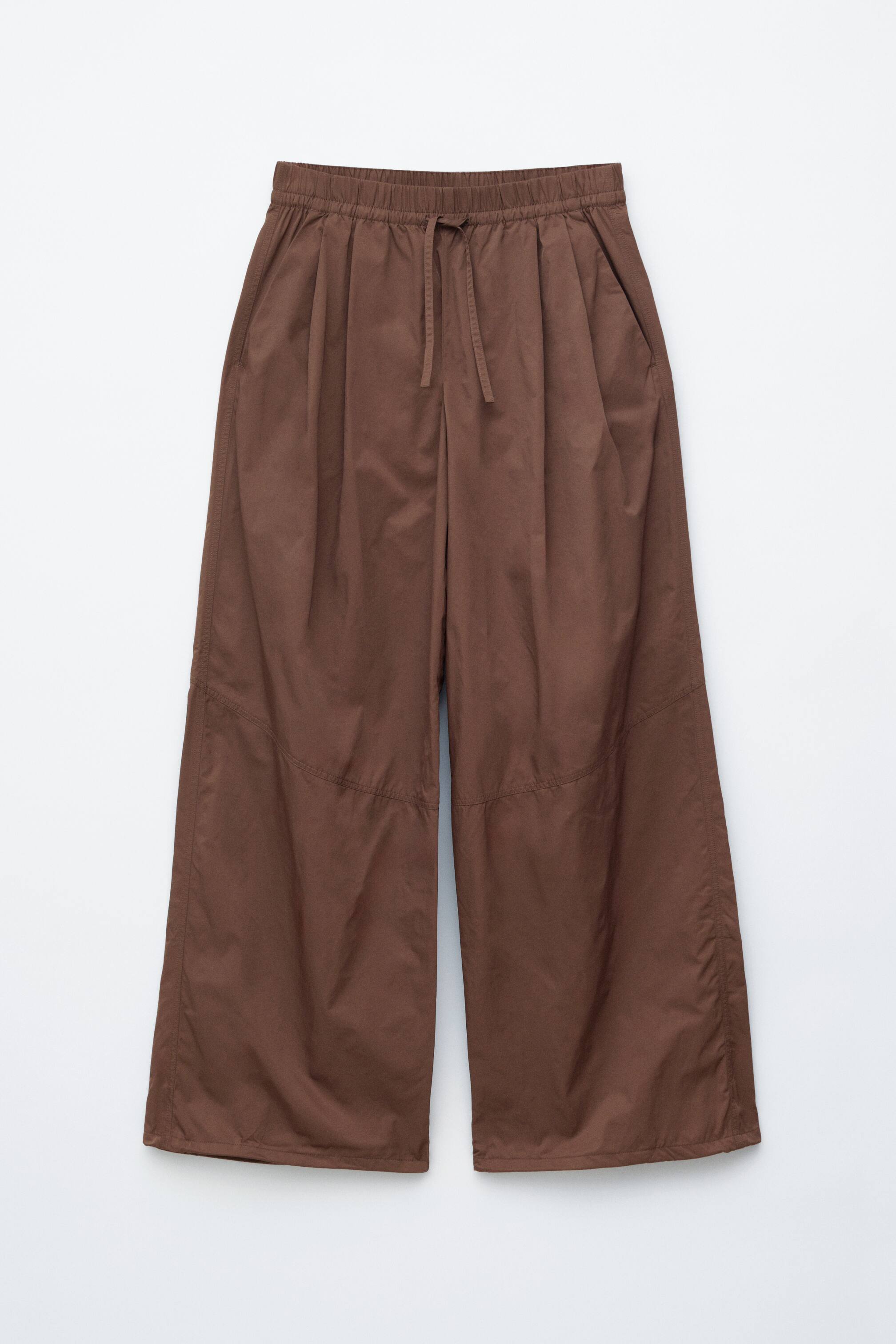 הצגת תמונה גדולה יותר: המכנסיים החומים מוצגים מקדימה, כוללים חגורת מותן אלסטית עם שרוך תואם וקפלים בולטים בחזית. תפרי חיתוך מפורטים נמשכים לאורך הרגליים, והמכנסיים מעוצבים בגזרה רחבה ורפויה עם שולי גימור נקיים.