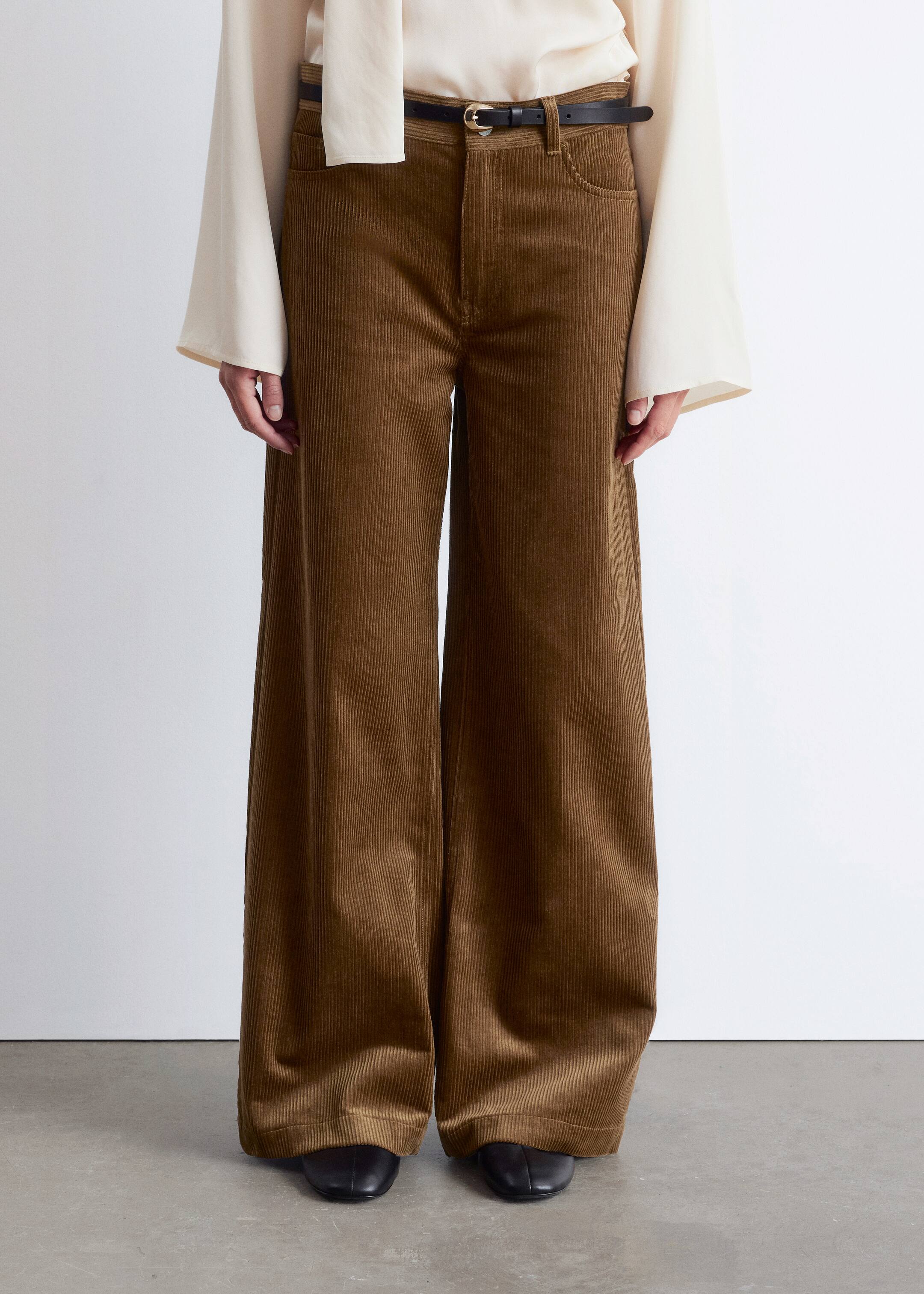 Agrandir l'image: Un pantalon en velours côtelé marron à jambes larges est présenté de face, avec une coupe taille haute et des côtes verticales distinctes, ainsi que des poches avant en forme de cuillère. Une ceinture noire avec une boucle dorée est portée à travers les passants de ceinture.