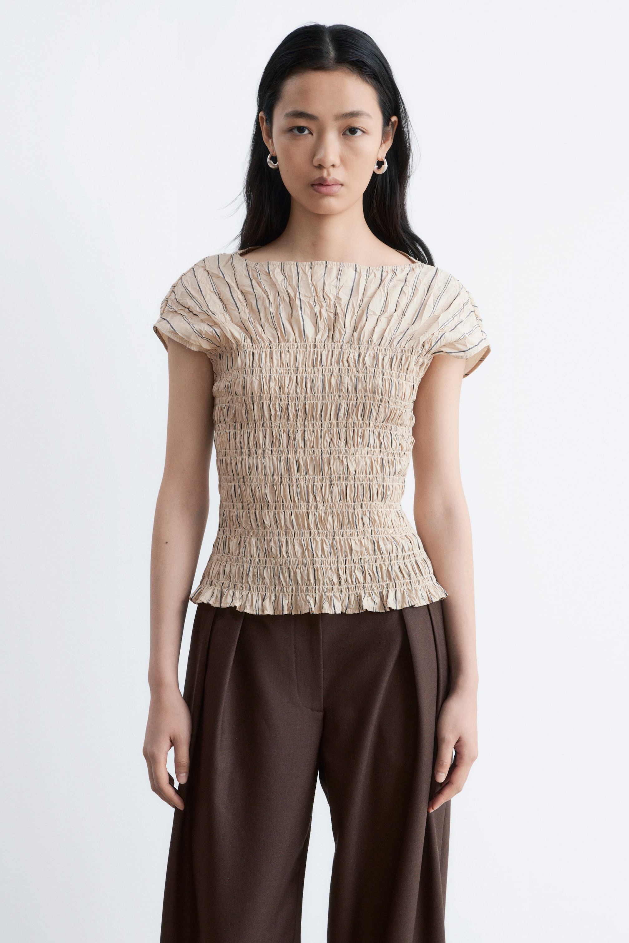 Ver imagem maior: Um top bege com efeito franzido é apresentado de frente, com um decote quadrado, mangas curtas estilo "cap" e uma textura enrugada com riscas verticais subtis ao longo do seu corpo ajustado e bainha com folhos. Está a ser combinado com calças largas de perna castanho-escuro que apresentam pregas frontais.