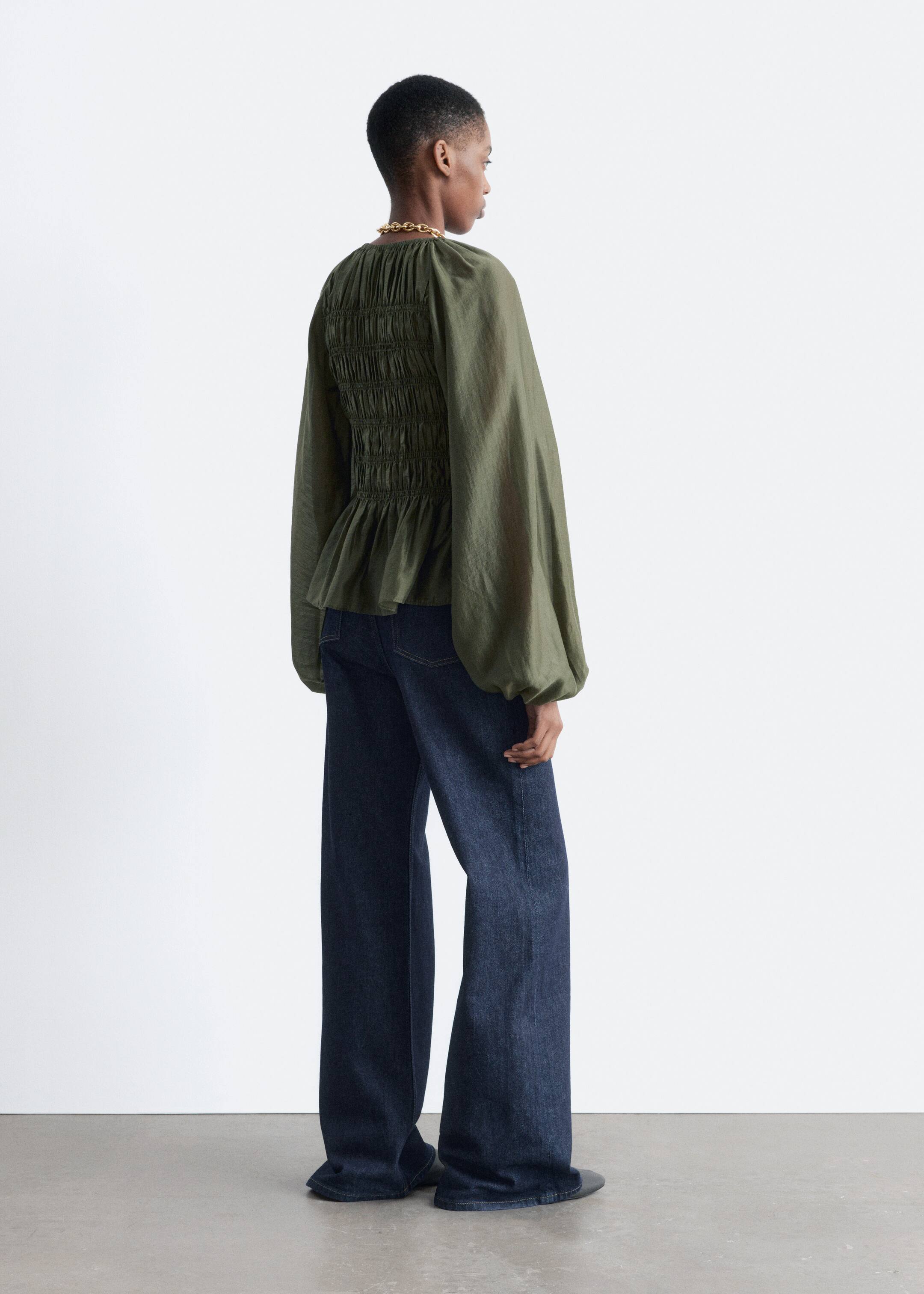 View larger image: A dark green top is shown from a rear-side angle, featuring extensive shirring across the upper back and waist that is flaring into a peplum hem. The top is designed with voluminous long sleeves that are gathering at the cuffs and is styled with dark blue, wide-leg jeans.