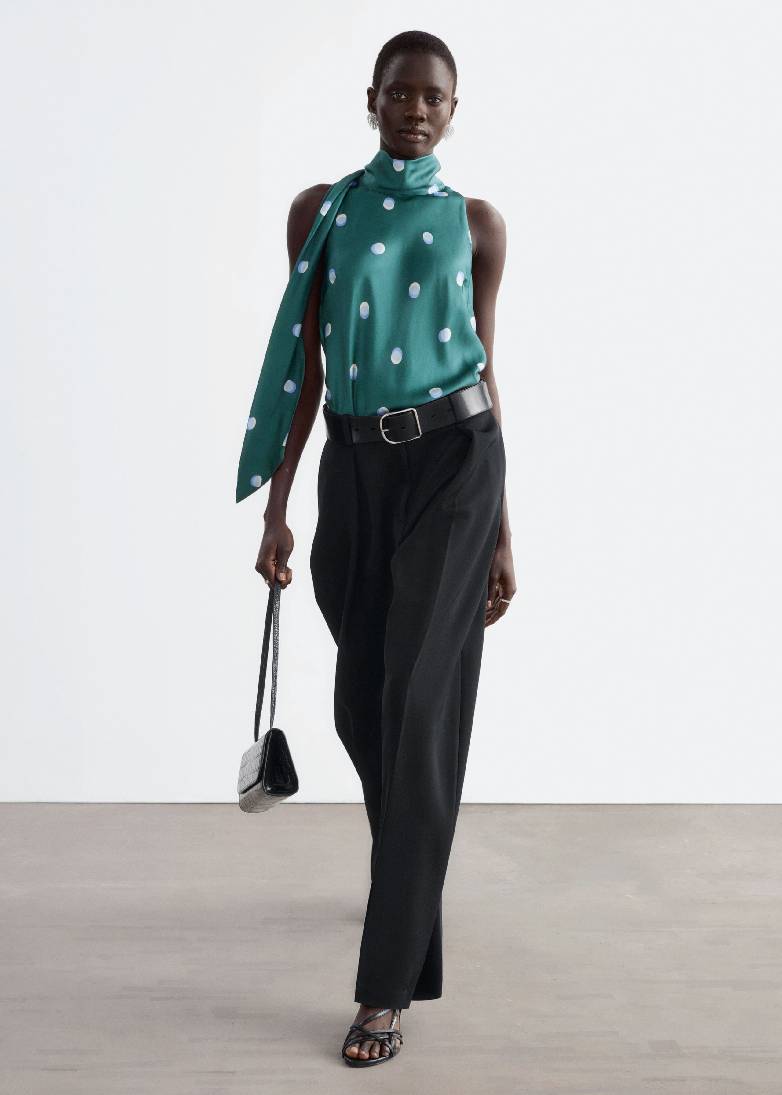 A green satin sleeveless top features a high-neck collar with an attached draped scarf and a blue and white polka dot pattern, worn by the model. The top is tucked into black wide-leg trousers styled with a black leather belt.