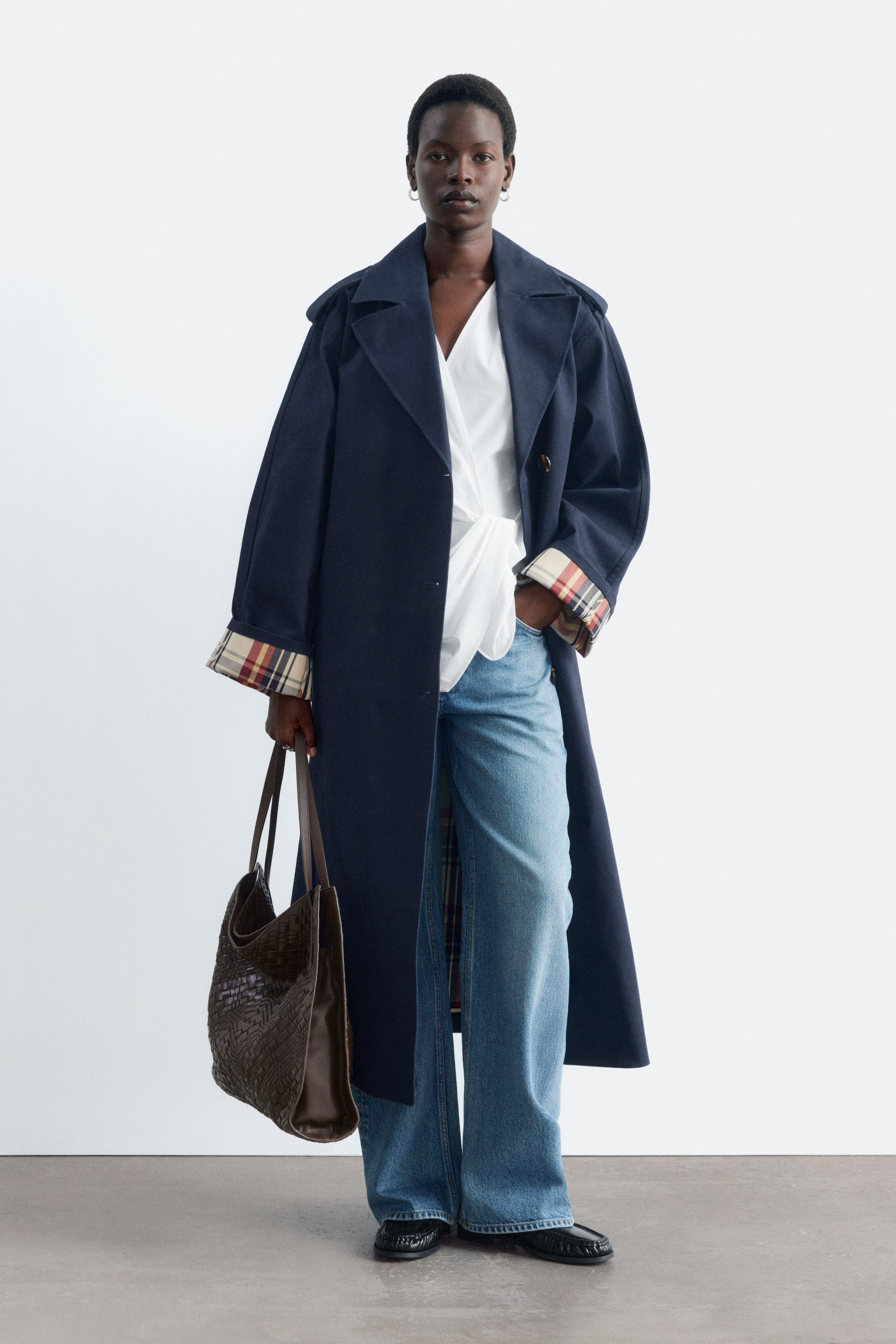 View larger image: A navy blue mid-length trench coat is worn open by the model, featuring wide lapels and folded cuffs that reveal a beige checked lining. The coat is layered over a white wrap top and wide-leg blue jeans, with a dark brown woven tote bag being held.