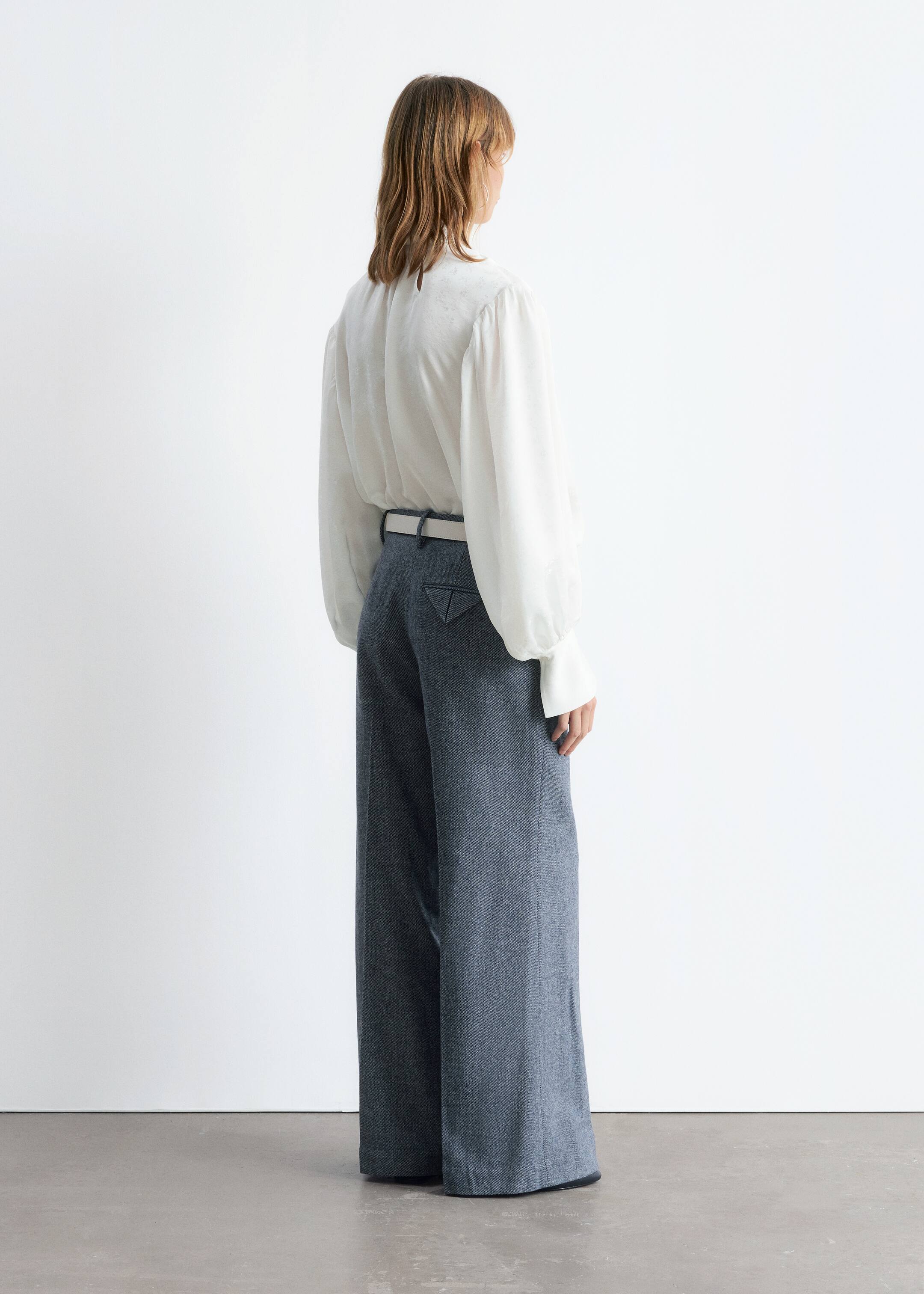 View larger image: A white, high-necked blouse with a subtle texture, featuring voluminous long sleeves and wide cuffs, is being worn tucked into high-waisted, wide-leg trousers. The blue melange trousers are featuring triangular back pockets and are cinched with a light-colored belt, all viewed from the back.
