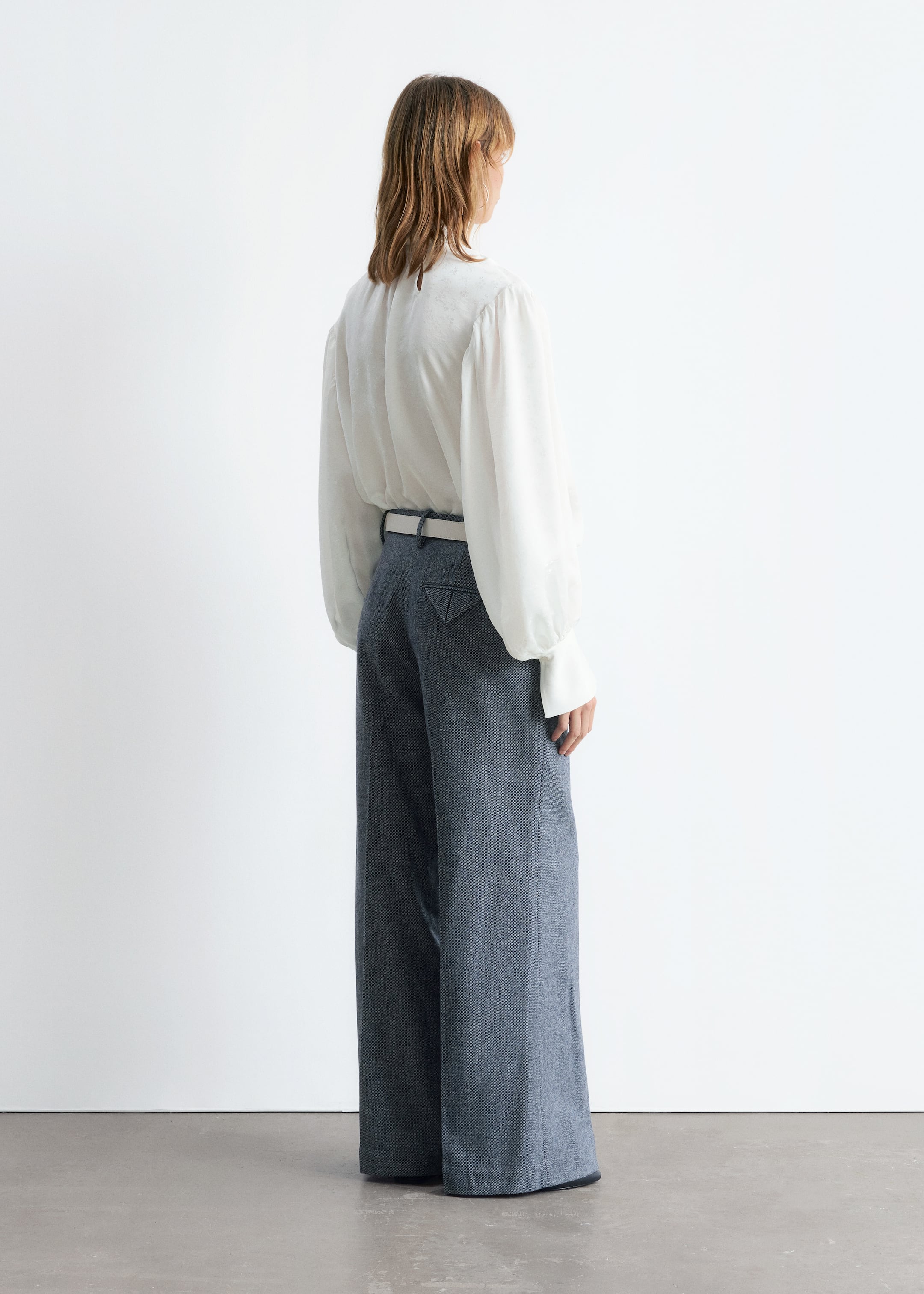 Agrandir l'image: Un chemisier blanc à col montant avec une texture subtile, doté de manches longues volumineuses et de larges poignets, est porté rentré dans un pantalon taille haute à jambes larges. Le pantalon bleu chiné présente des poches arrière triangulaires et est ceinturé avec une ceinture de couleur claire, le tout vu de dos.